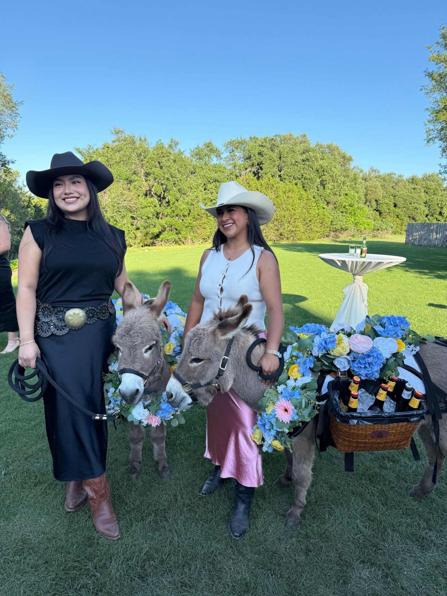 Two women stand outdoors with a table of flowers, trees, and a clear blue sky in the background.