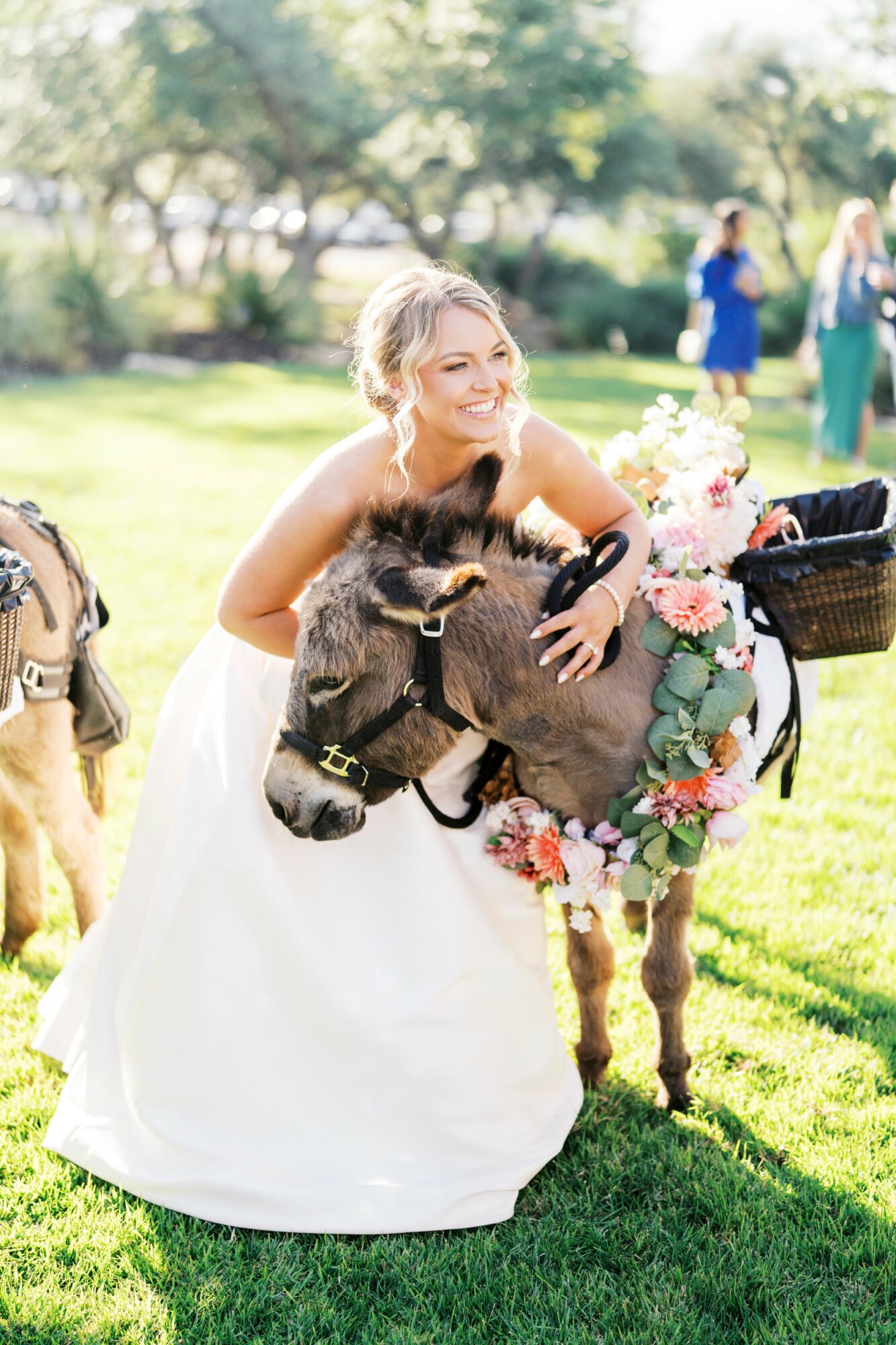 Bride in wedding dress hugging a small pony decorated with flowers outdoors, with people and trees in background.
