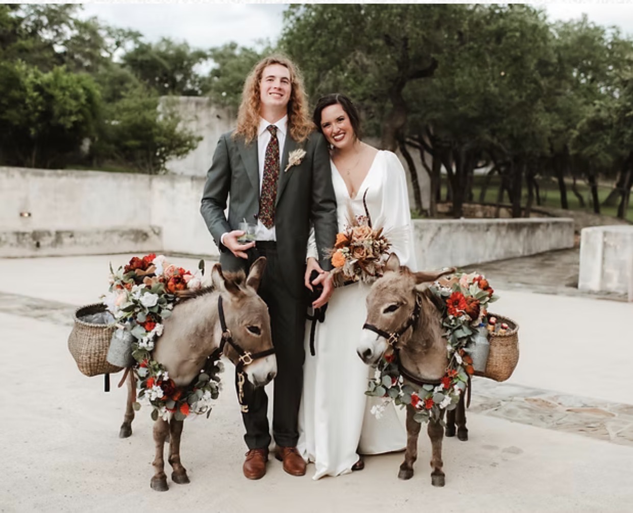 A couple in wedding attire standing outdoors with two donkeys decorated with flowers and baskets, trees in the background.
