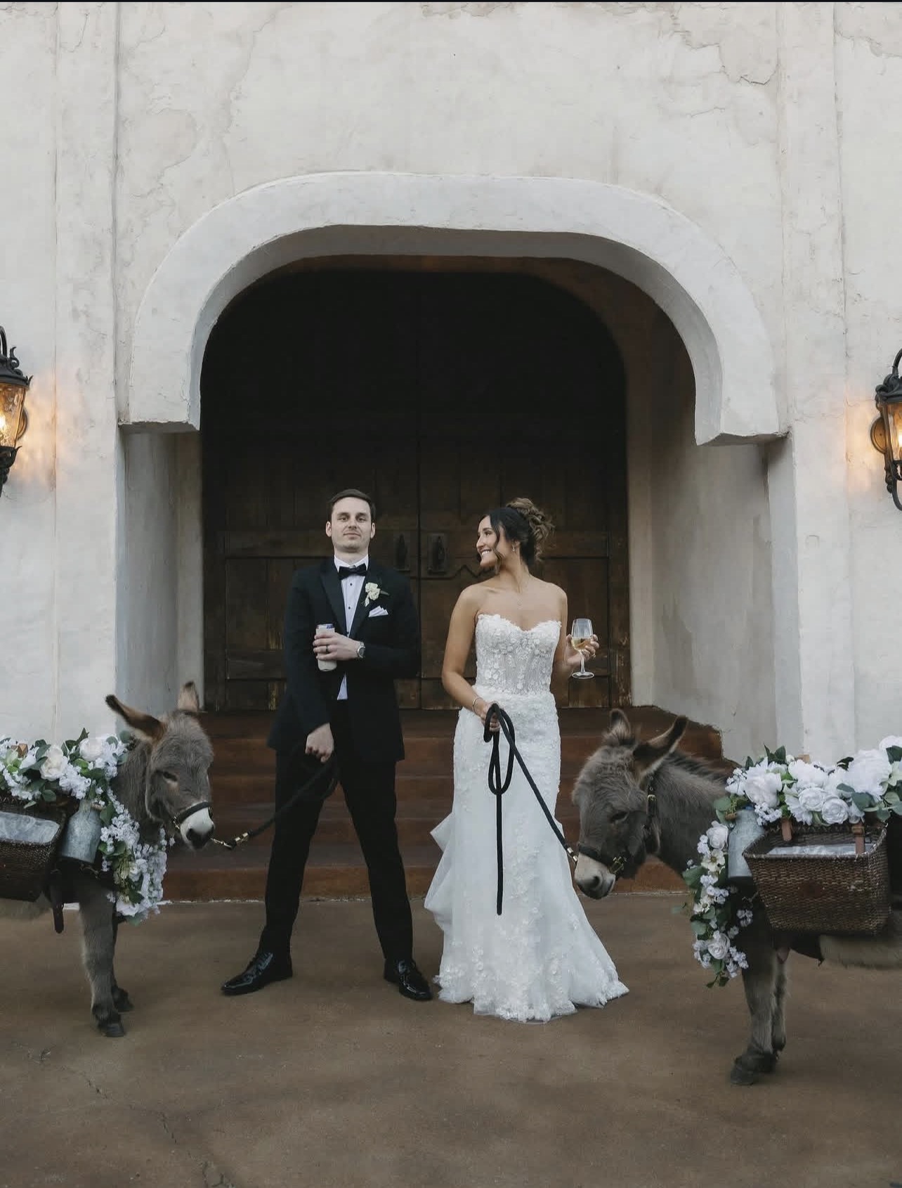 Bride and groom stand in front of a large wooden door, surrounded by decorated reindeer figures and floral arrangements.