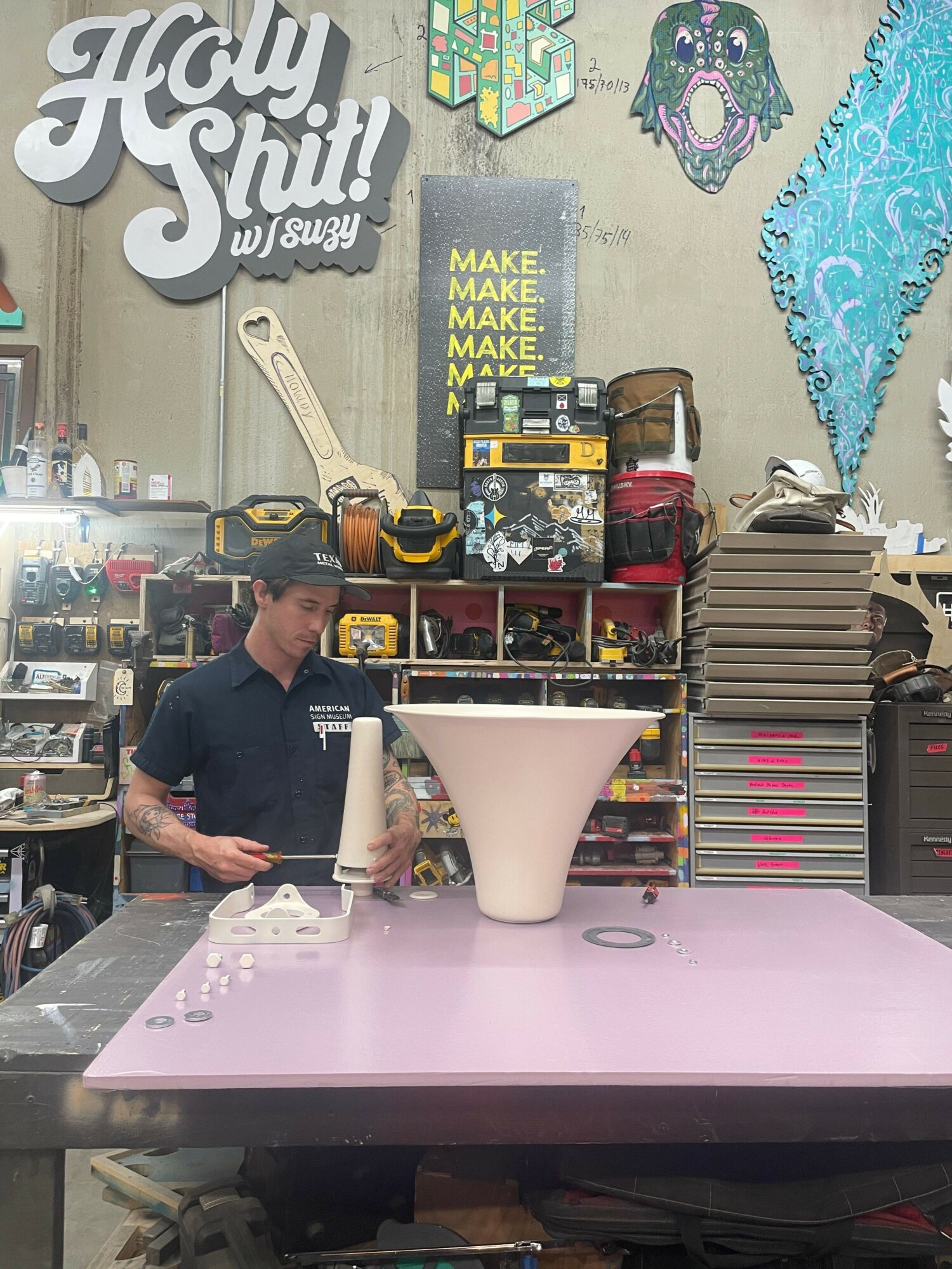 A person working at a cluttered table with various objects, shelves, and signs in the background, viewed from the side.