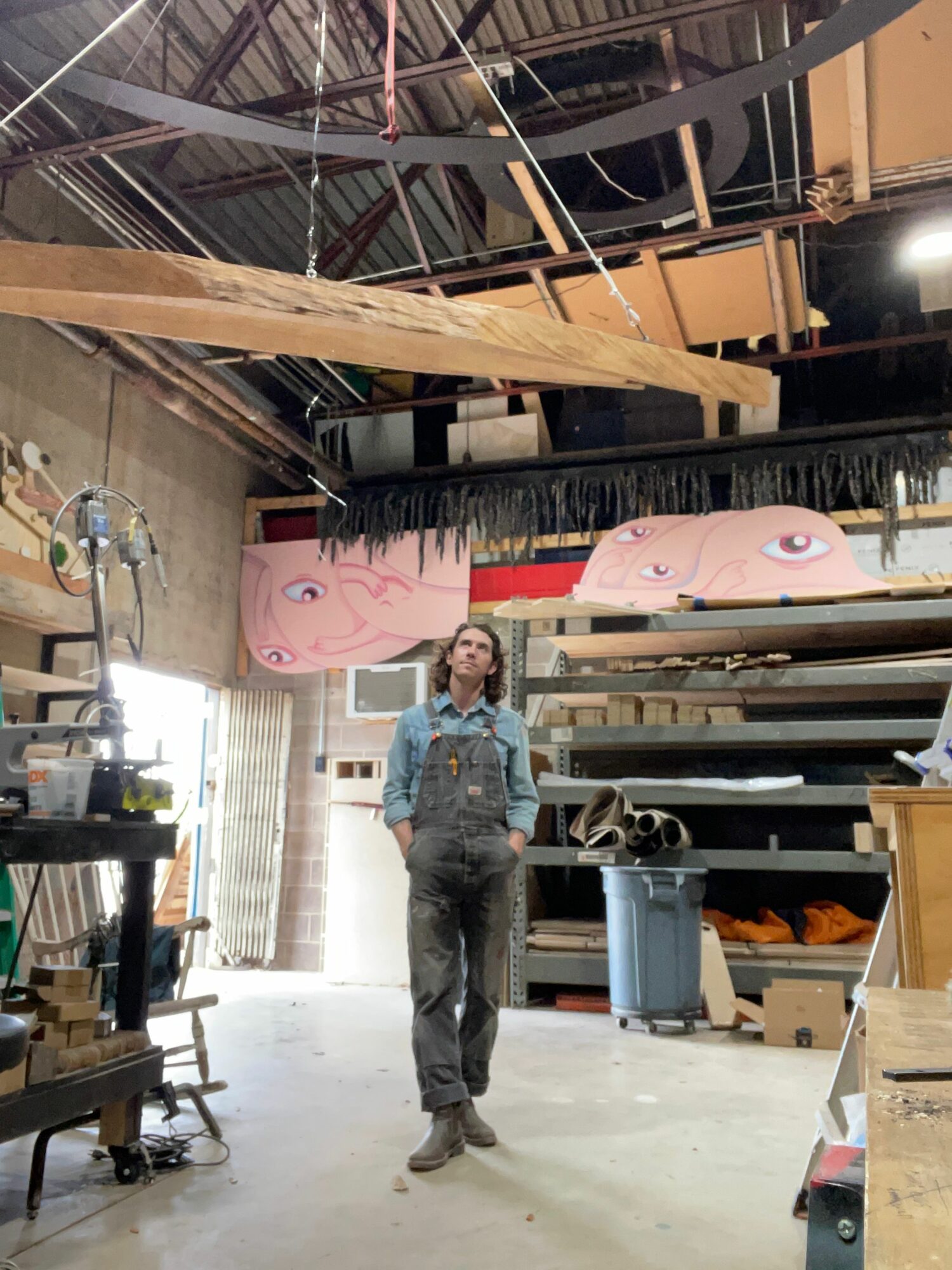 Person standing in a workshop with wooden beams, tools, and pink pig decorations in the background.