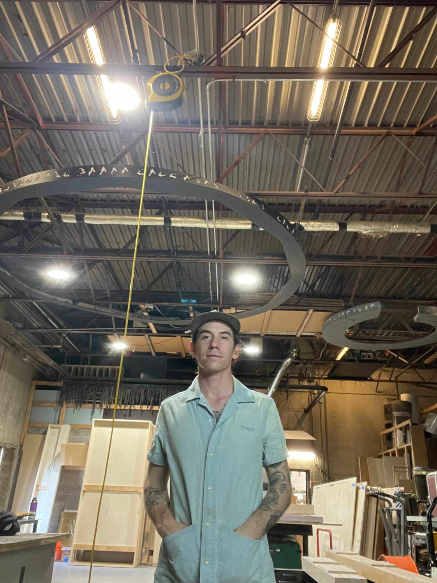Person in a light blue shirt standing in a workshop with equipment and tools, ceiling with exposed beams and lighting.
