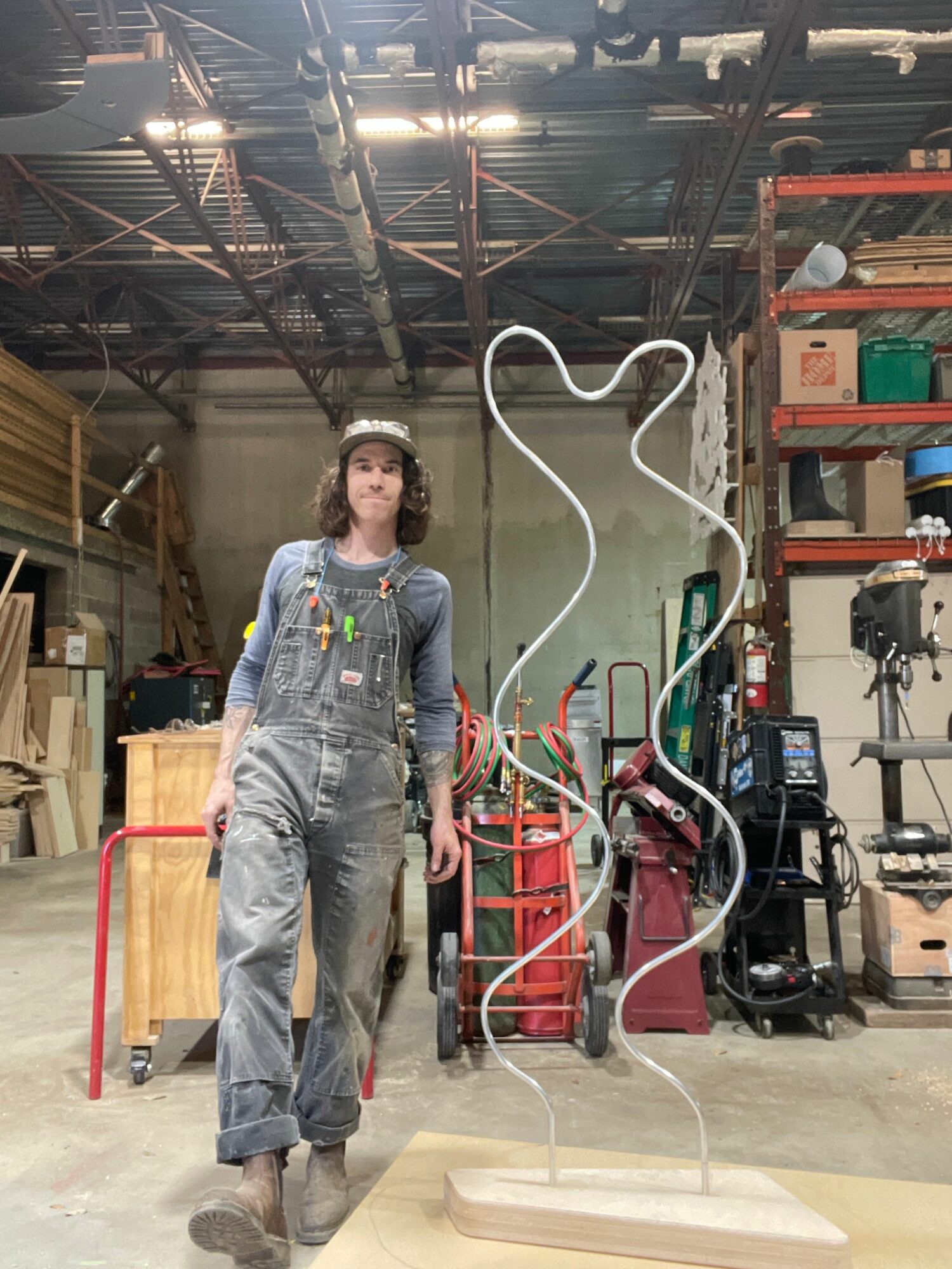 Person in gray coveralls standing in industrial workshop with equipment and tools, facing camera, with ceiling and machinery overhead.