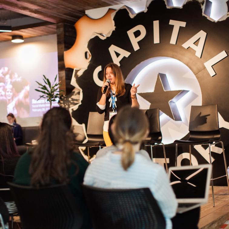 Woman speaking on stage in front of a large 'CAPITAL' sign with a star, audience seated facing her, indoor setting.