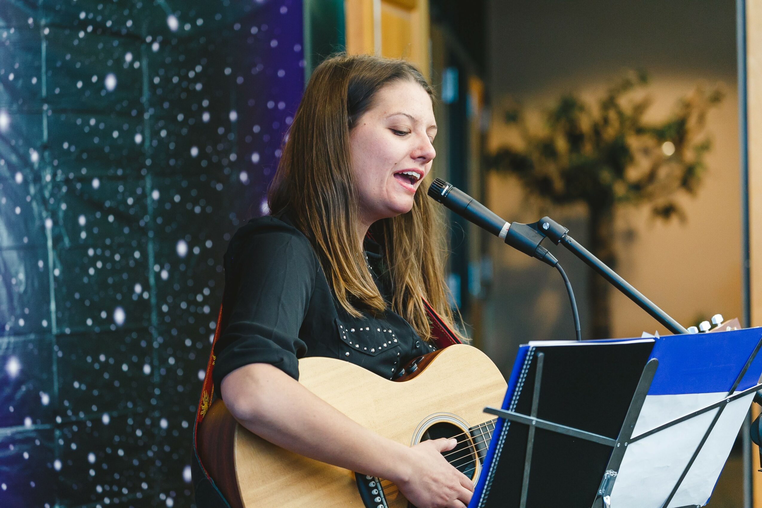 Woman singing into a microphone while playing an acoustic guitar, with a music stand in front of her, in a decorated indoor setting.