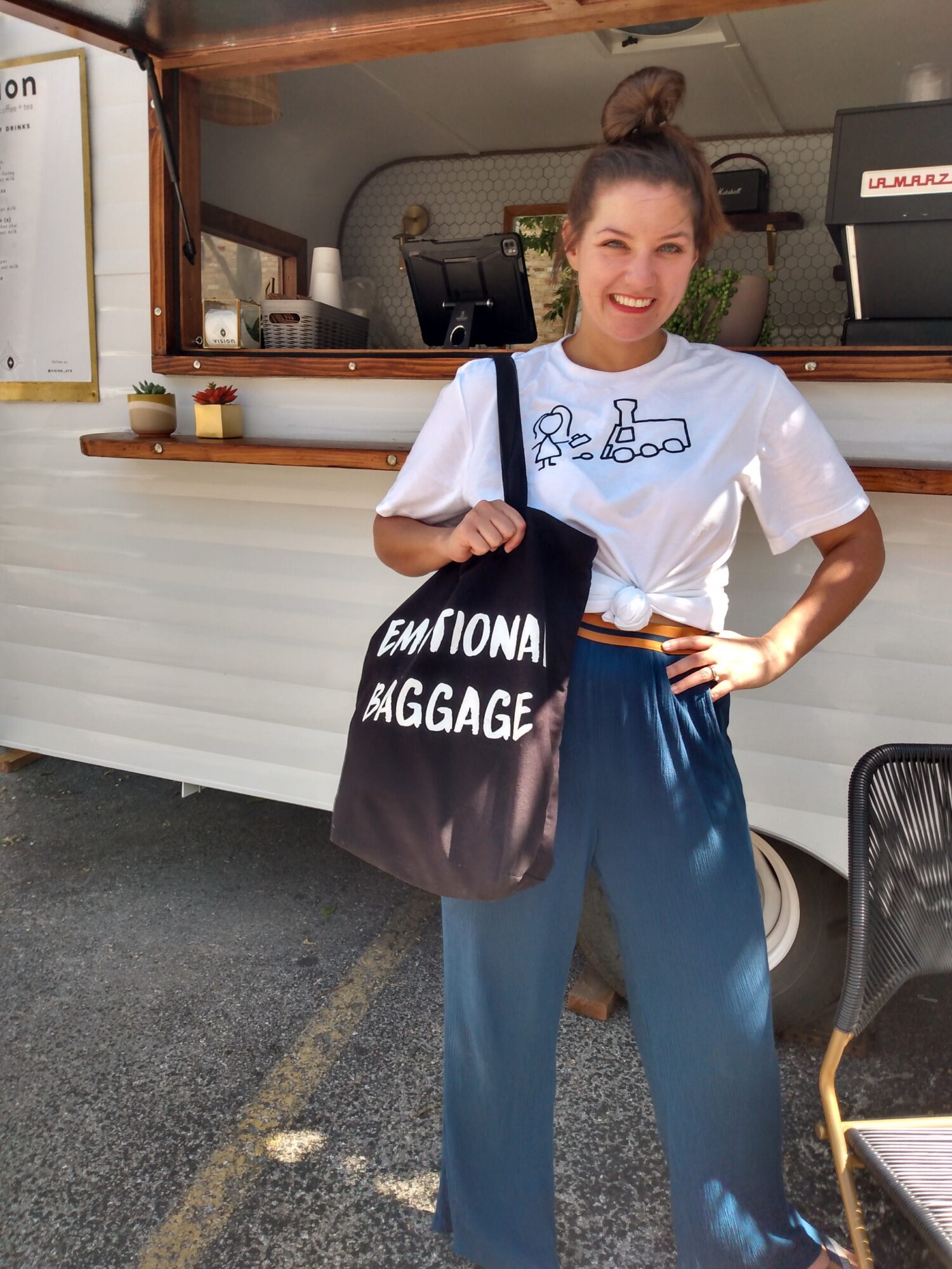 Smiling woman with a bun hairstyle, wearing a white t-shirt and blue pants, holding a black bag with white text, standing outside a food truck.