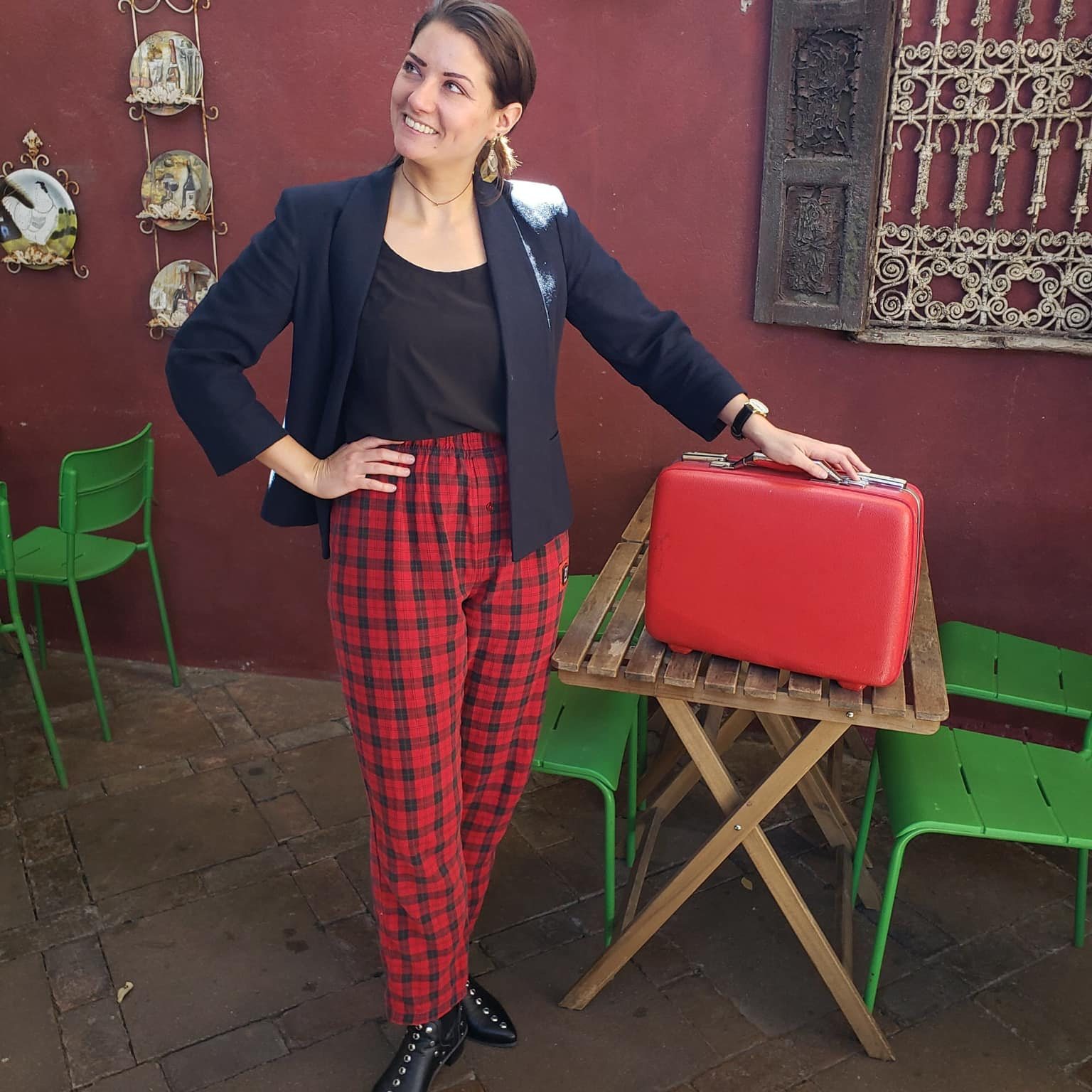 Woman standing indoors, smiling, wearing a black blazer and red plaid pants, with a red suitcase on a wooden table.