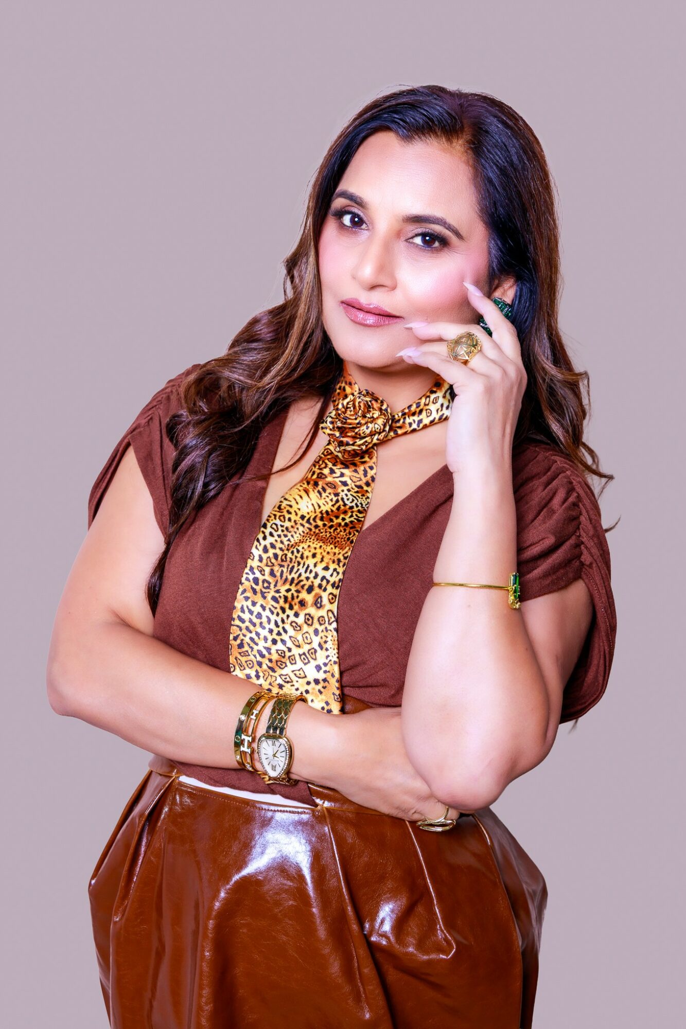 Woman with long hair, wearing a brown top and shiny brown skirt, holding a phone to her ear, looking at the camera.