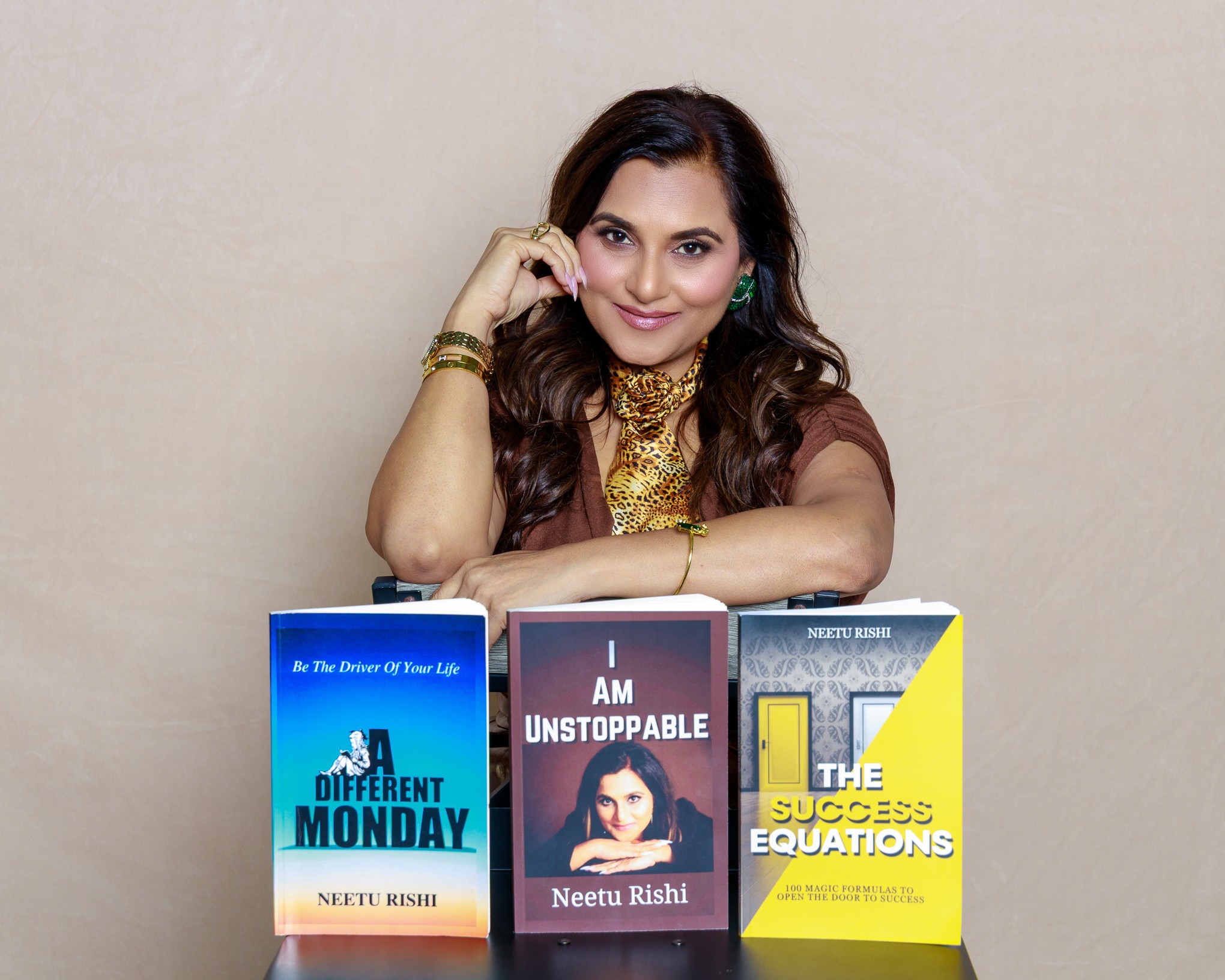 Woman with long dark hair, wearing gold jewelry, sitting behind three books on a table, smiling at the camera.