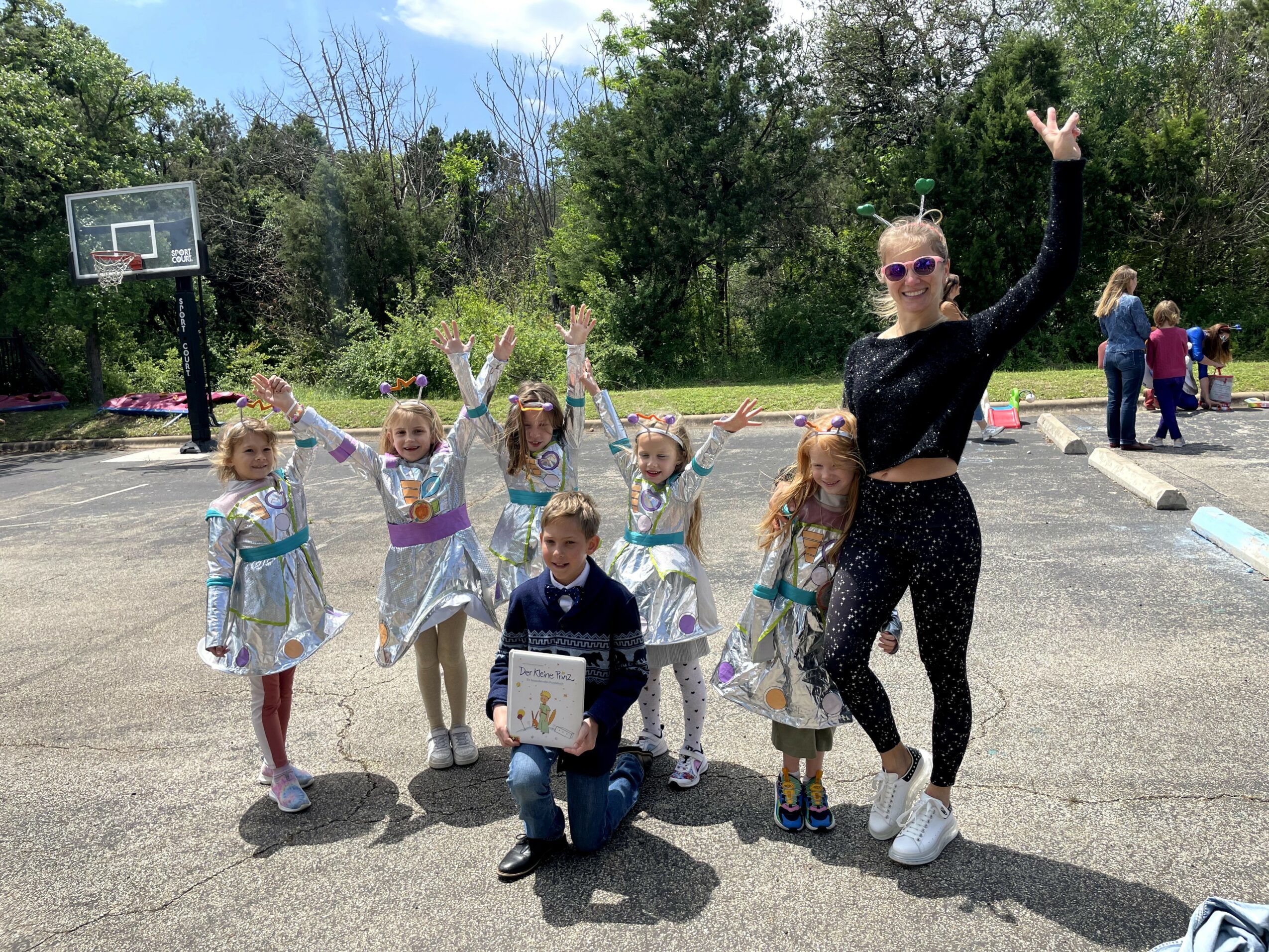 Group of children and an adult posing outdoors on a basketball court, some raising hands, with trees and people in background.