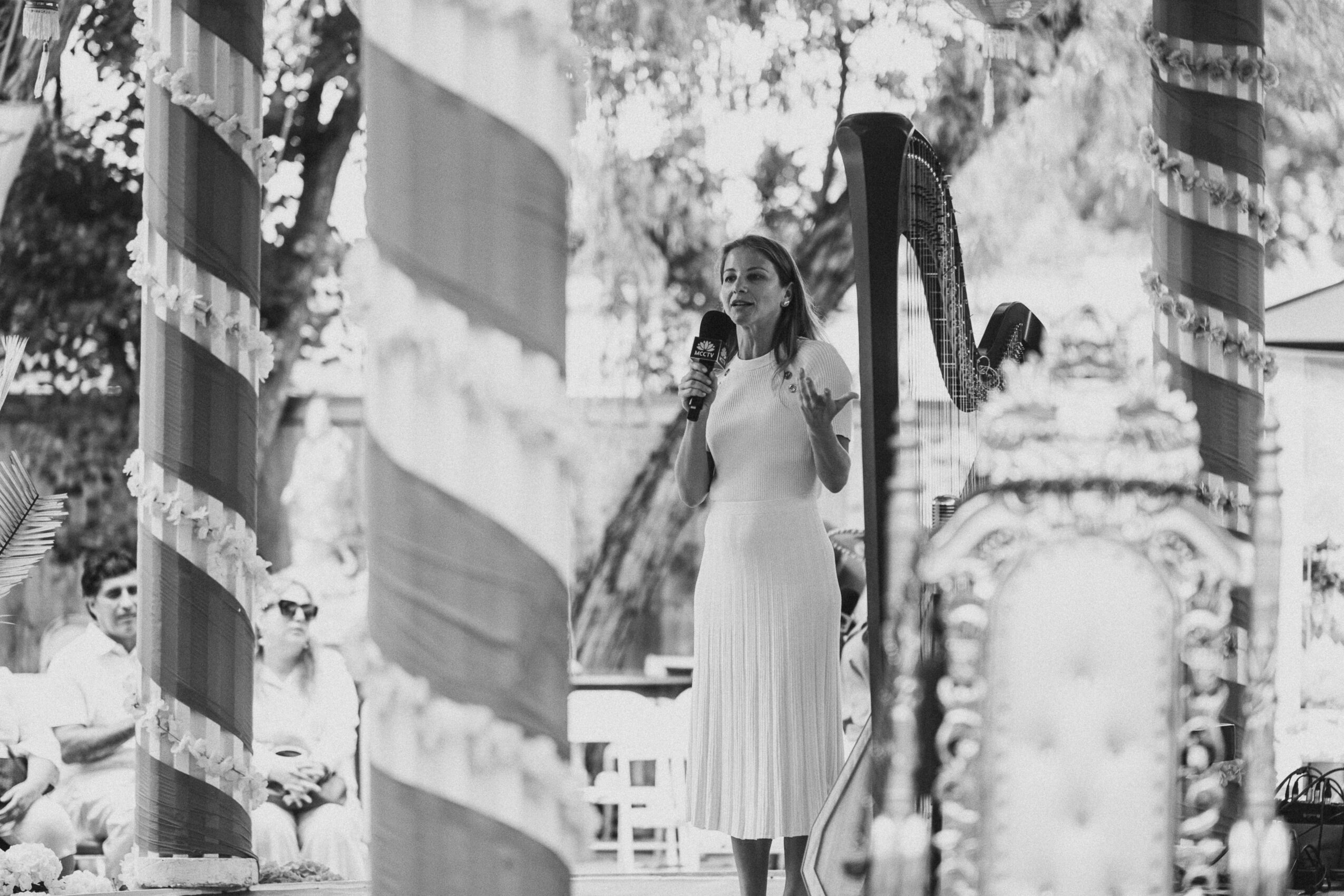 A woman in a long dress speaks into a microphone on an outdoor stage with decorative columns and trees in the background.