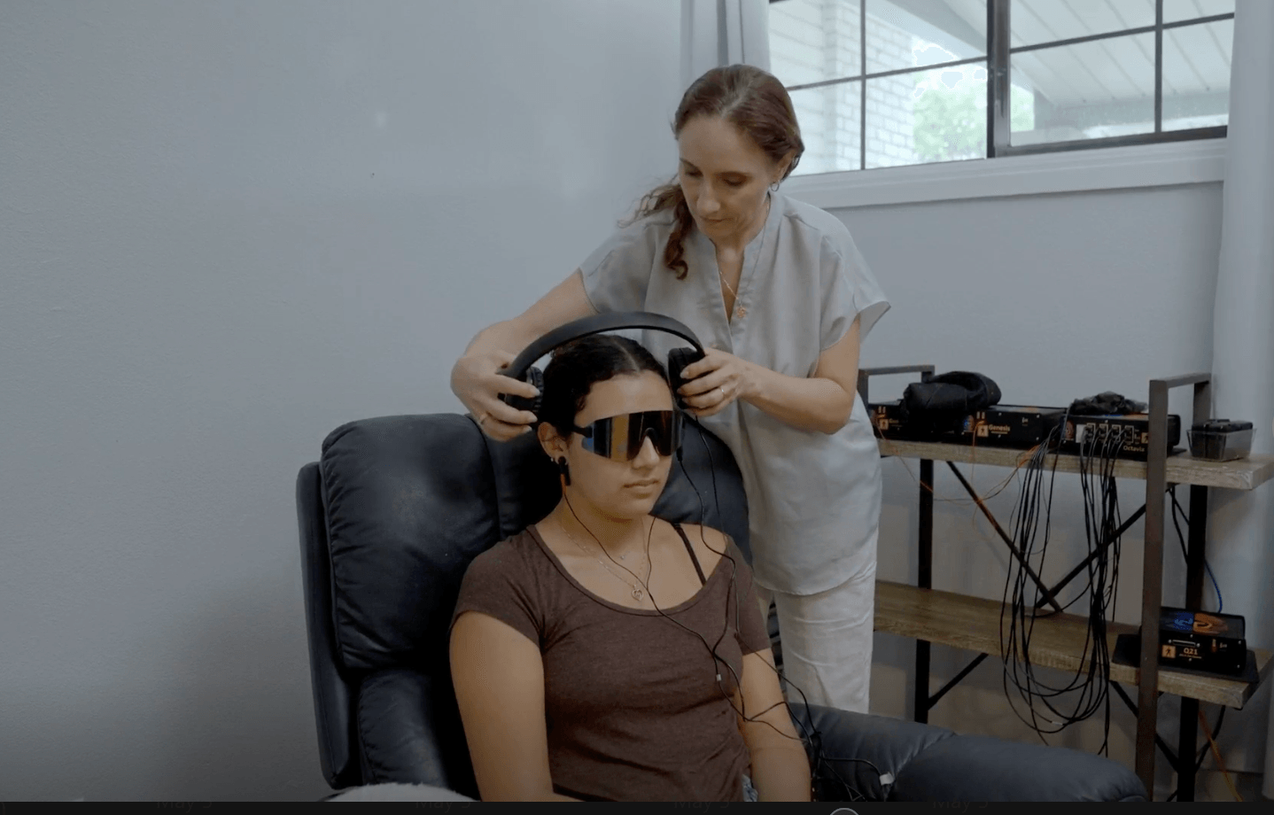 A woman wearing glasses is seated with a device on her head, while a technician adjusts equipment in a room with a window.