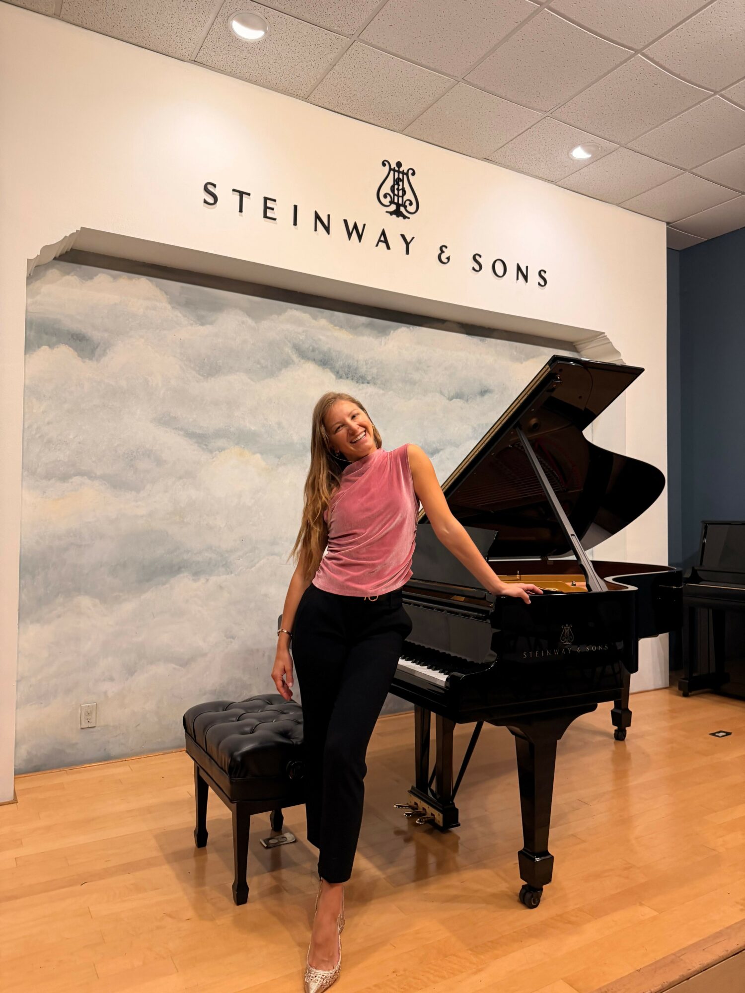 Woman in pink shirt and black pants standing next to a grand piano in a room with a Steinway & Sons sign on the wall.