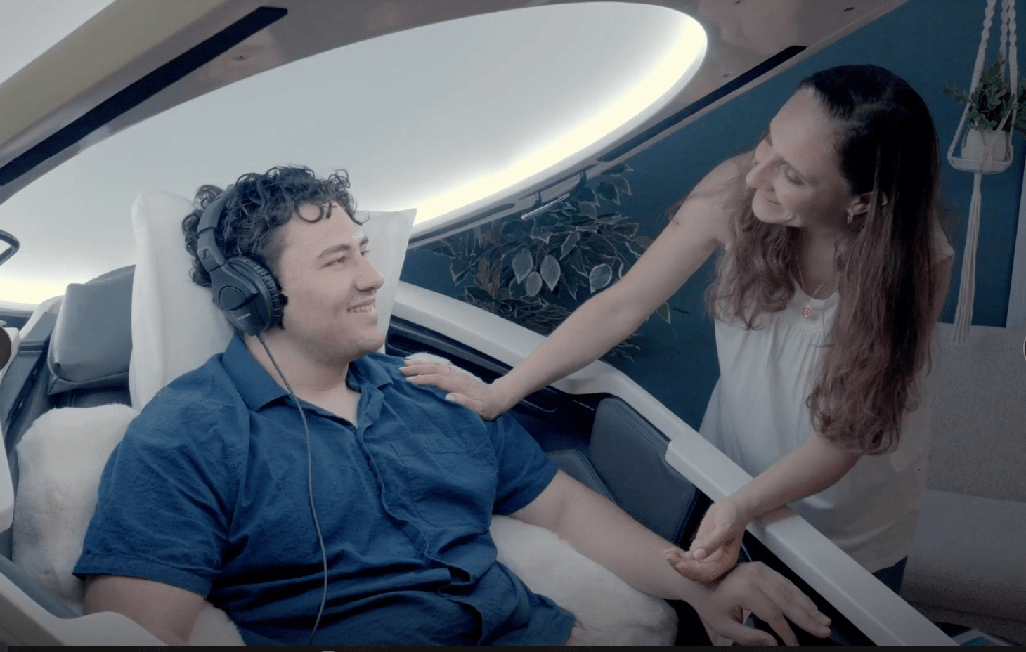 A woman interacts with a man wearing headphones in a futuristic setting, with a curved white structure and plants in the background.