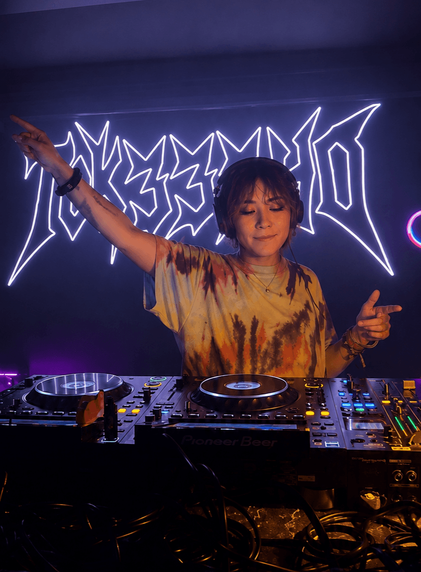 Female DJ with short hair and tie-dye shirt performing at a nightclub with neon sign background