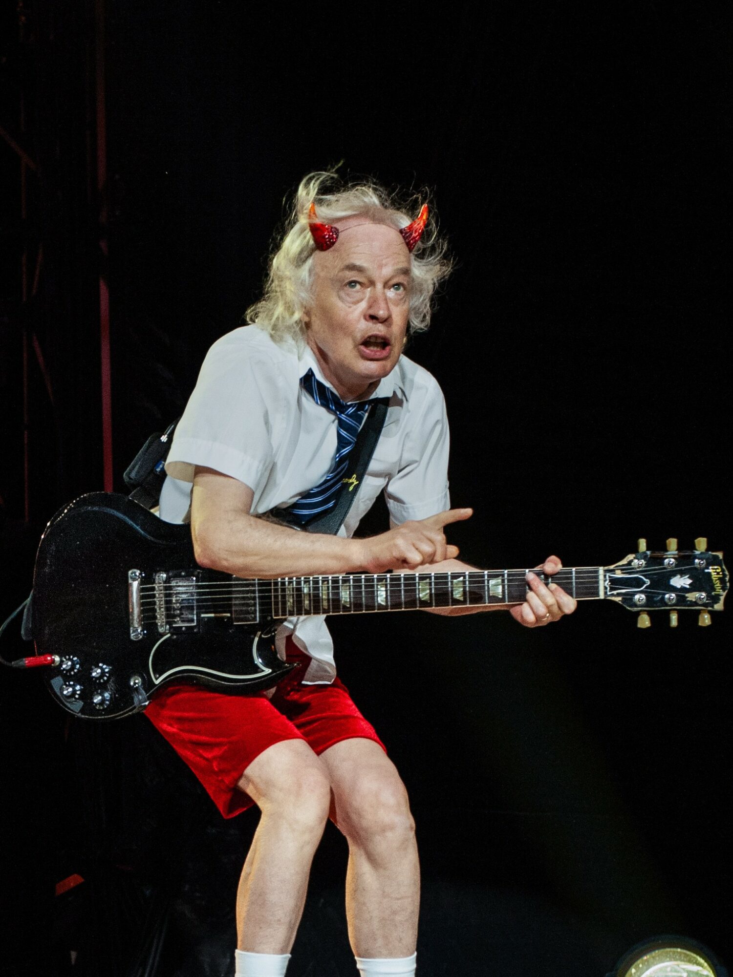 Person with blonde hair, red headband, white shirt, blue tie, red shorts, holding a black electric guitar, on stage.