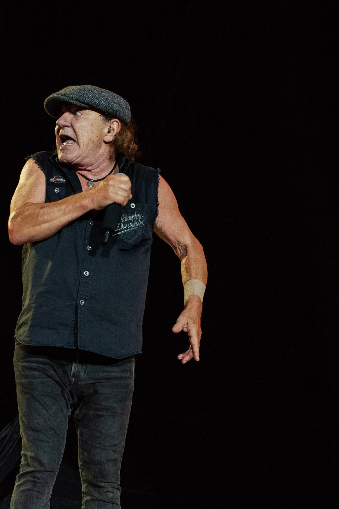 Person wearing a hat and sleeveless shirt, gesturing with one arm, against a dark background.