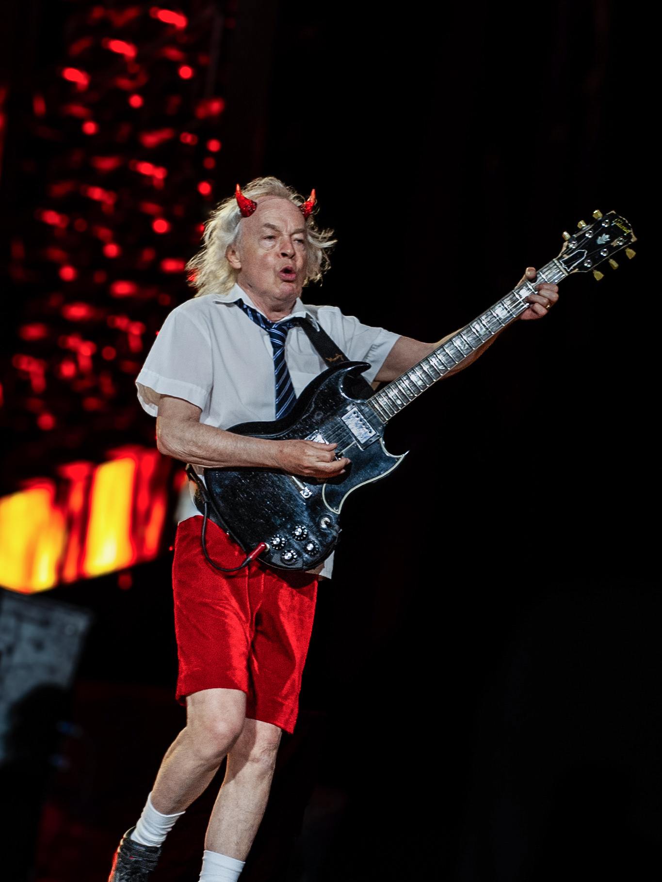 Older man with gray hair, devil horns, white shirt, red shorts, playing electric guitar on stage.