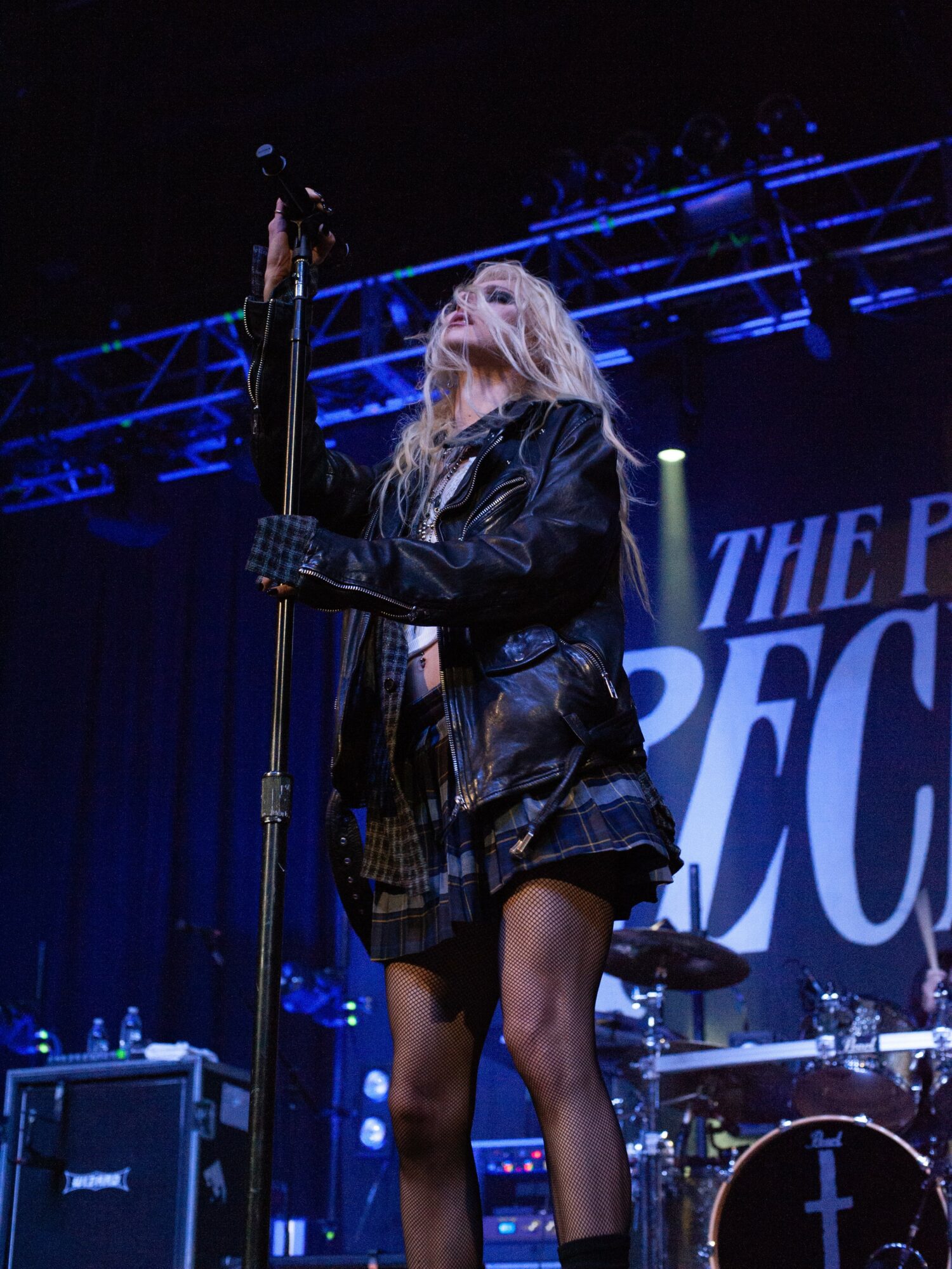 Female singer with long blonde hair performs on stage, holding a microphone, wearing a black leather jacket and skirt.