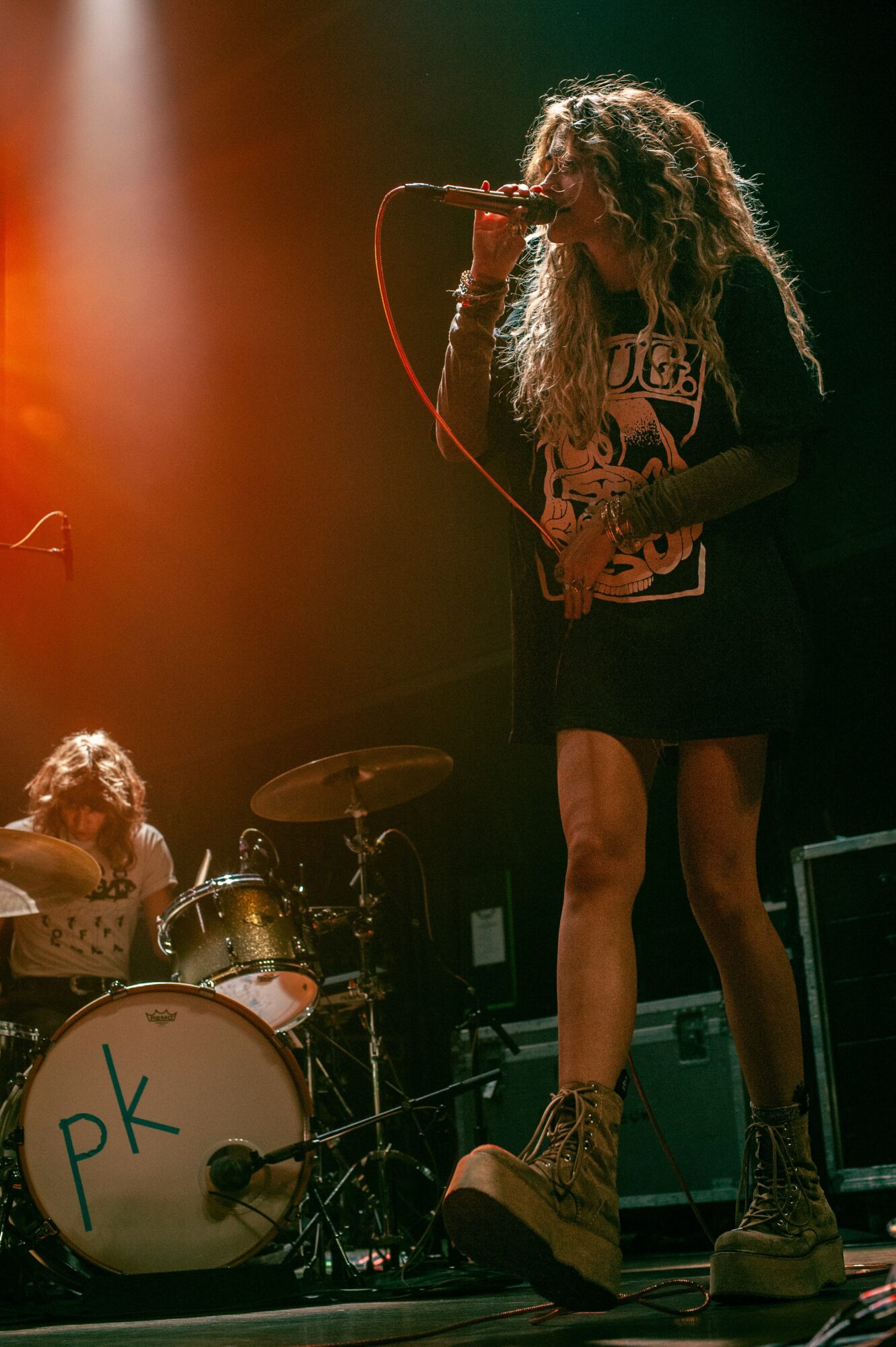 A female singer with curly hair performs on stage, holding a microphone, with a drummer in the background, stage lighting present.