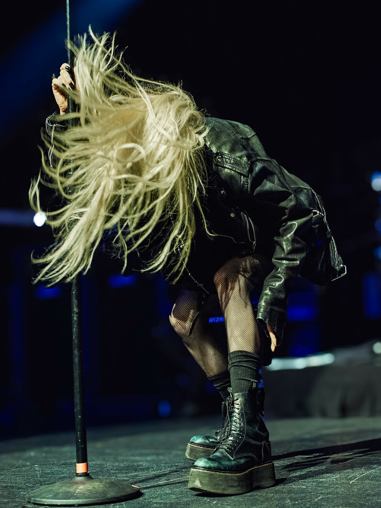 Person with long blonde hair bending forward on stage, wearing a black leather jacket and platform boots, dark background.