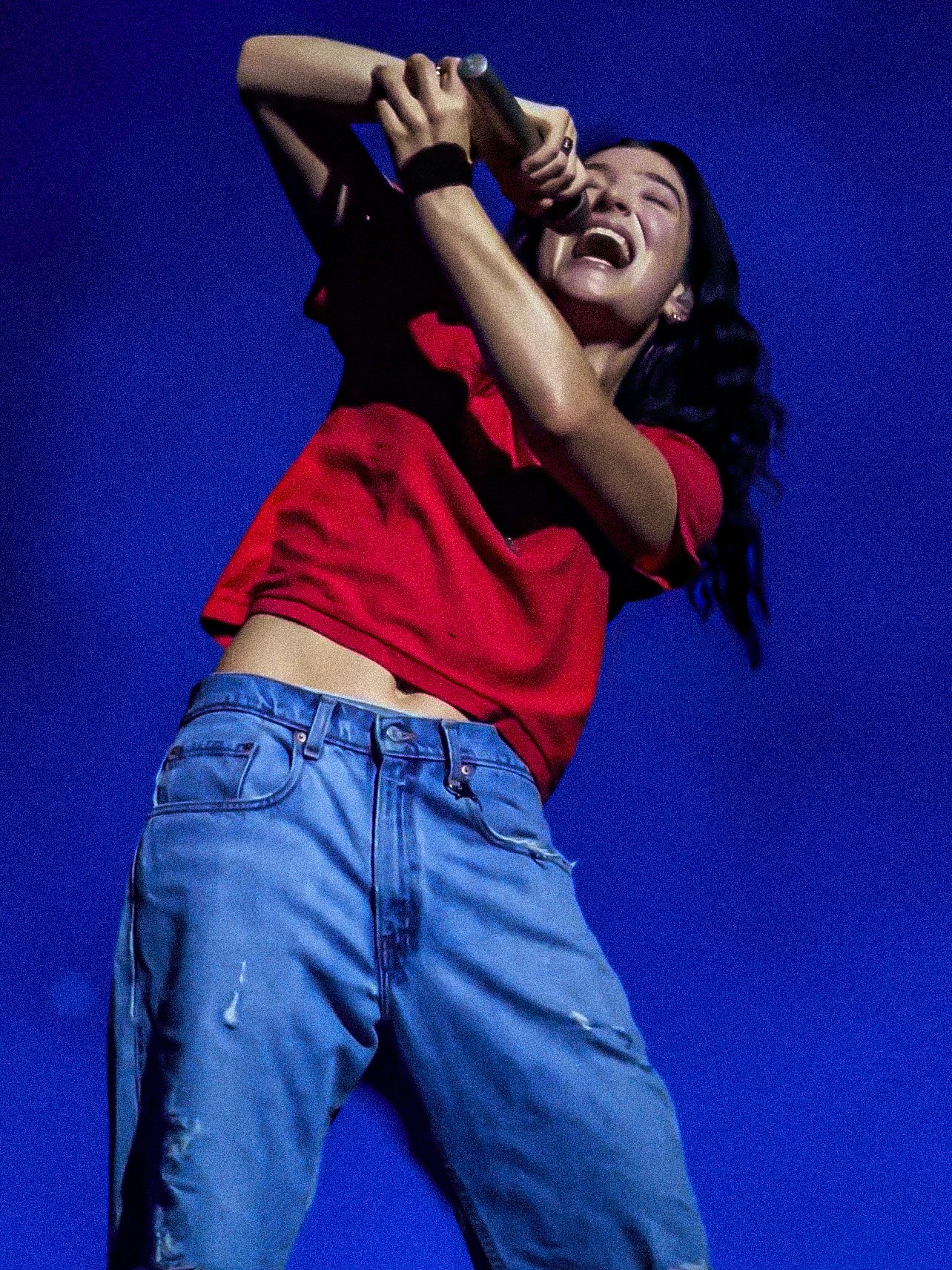 Person laughing, wearing a red shirt and blue jeans, holding a camera or phone, against a dark blue background.