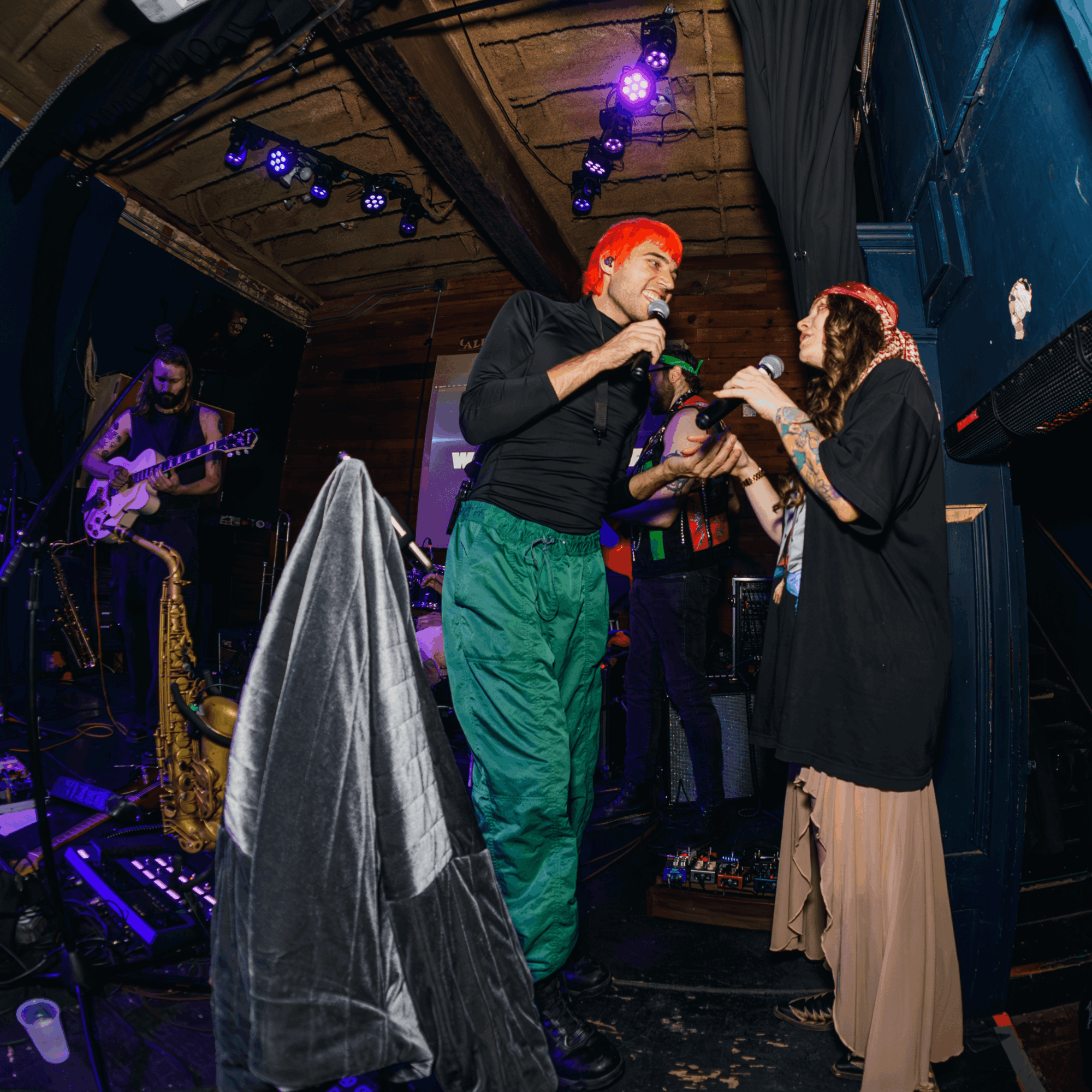 Two women are on stage, one with bright red hair and the other with a headband, engaging in a conversation. A musician plays guitar in the background. Stage lights illuminate the scene.