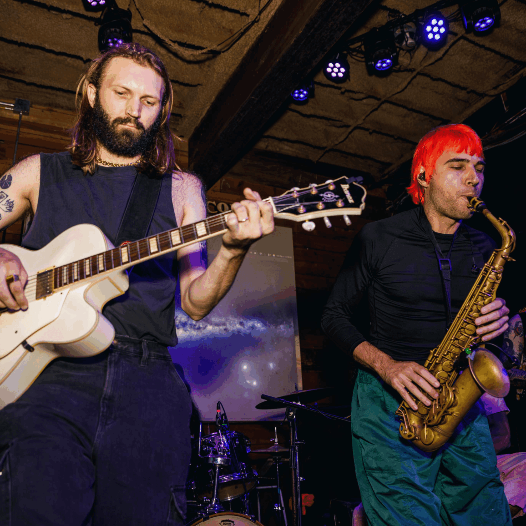Two musicians perform on stage, one playing guitar and the other playing saxophone, with stage lighting overhead.
