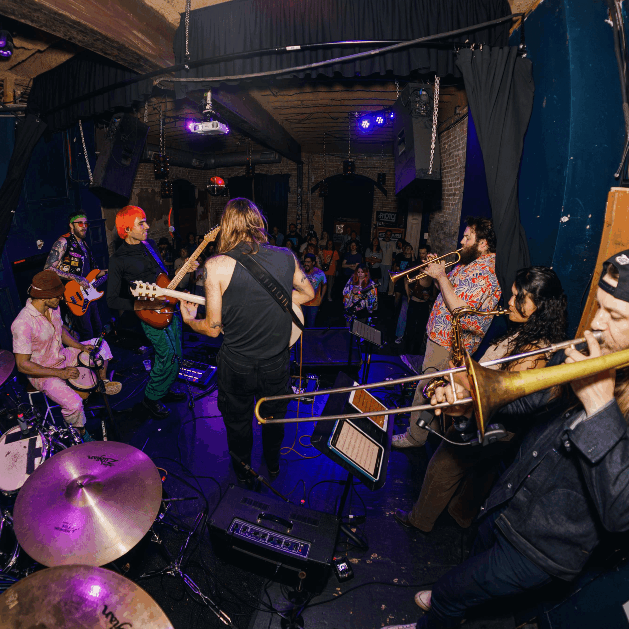 Band performs live music on stage with audience in a dimly lit venue, some playing instruments, others watching.