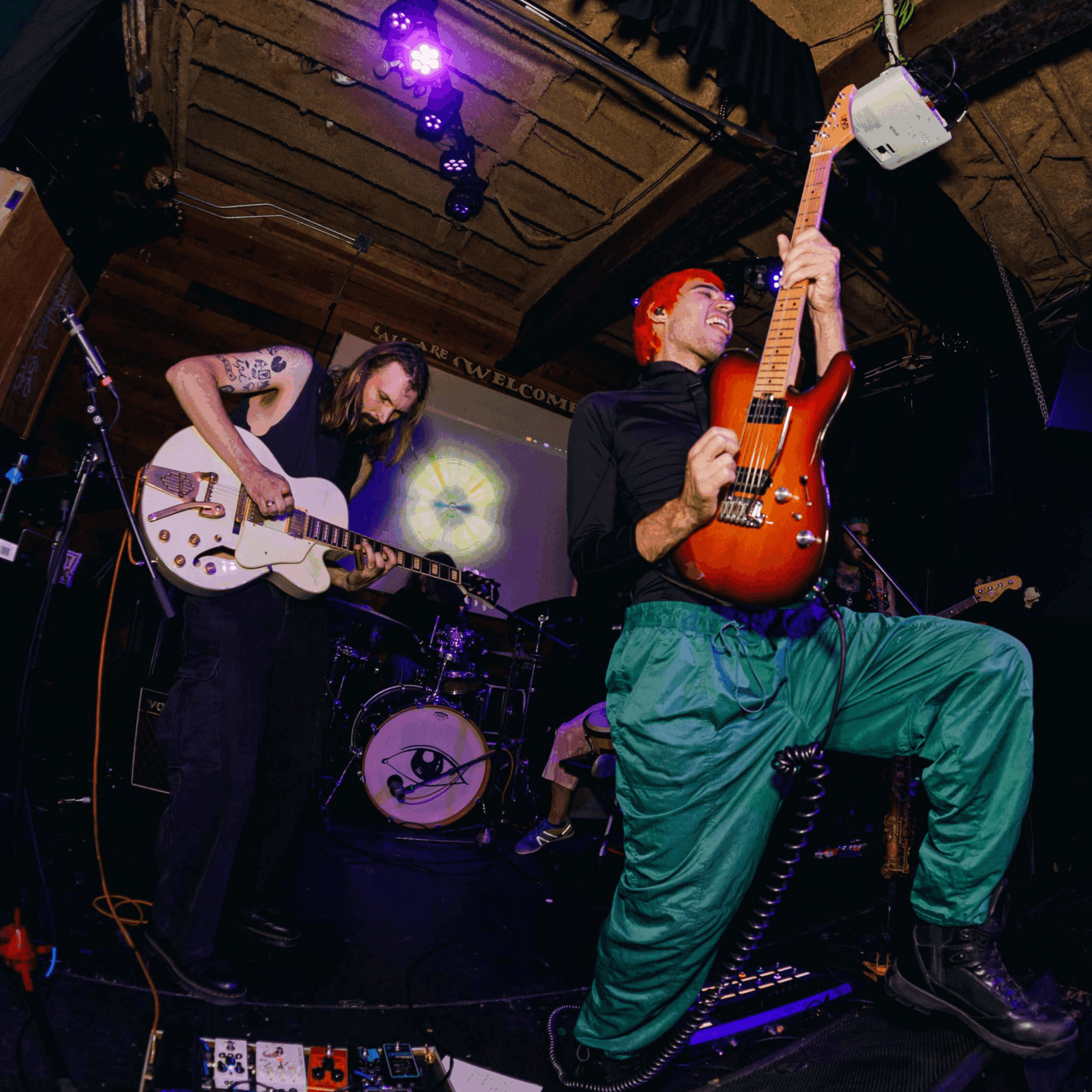 Two musicians perform on stage, one playing guitar and the other with an electric guitar, in a dimly lit venue.