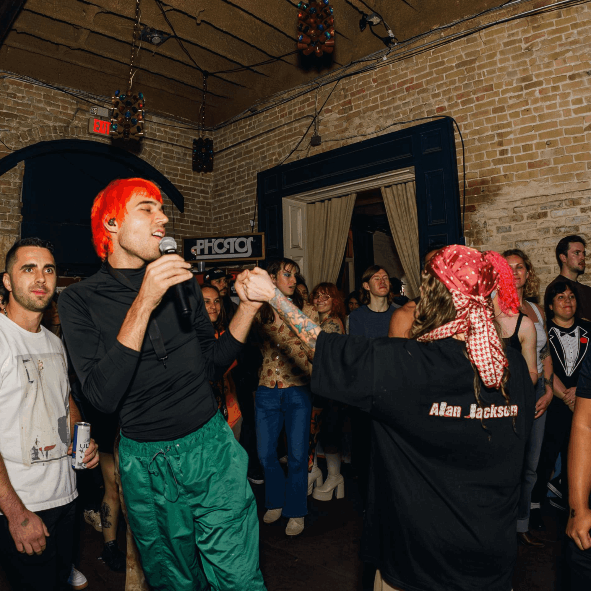 Person with bright red hair singing into a microphone while dancing with a woman in a red headscarf, surrounded by a crowd.