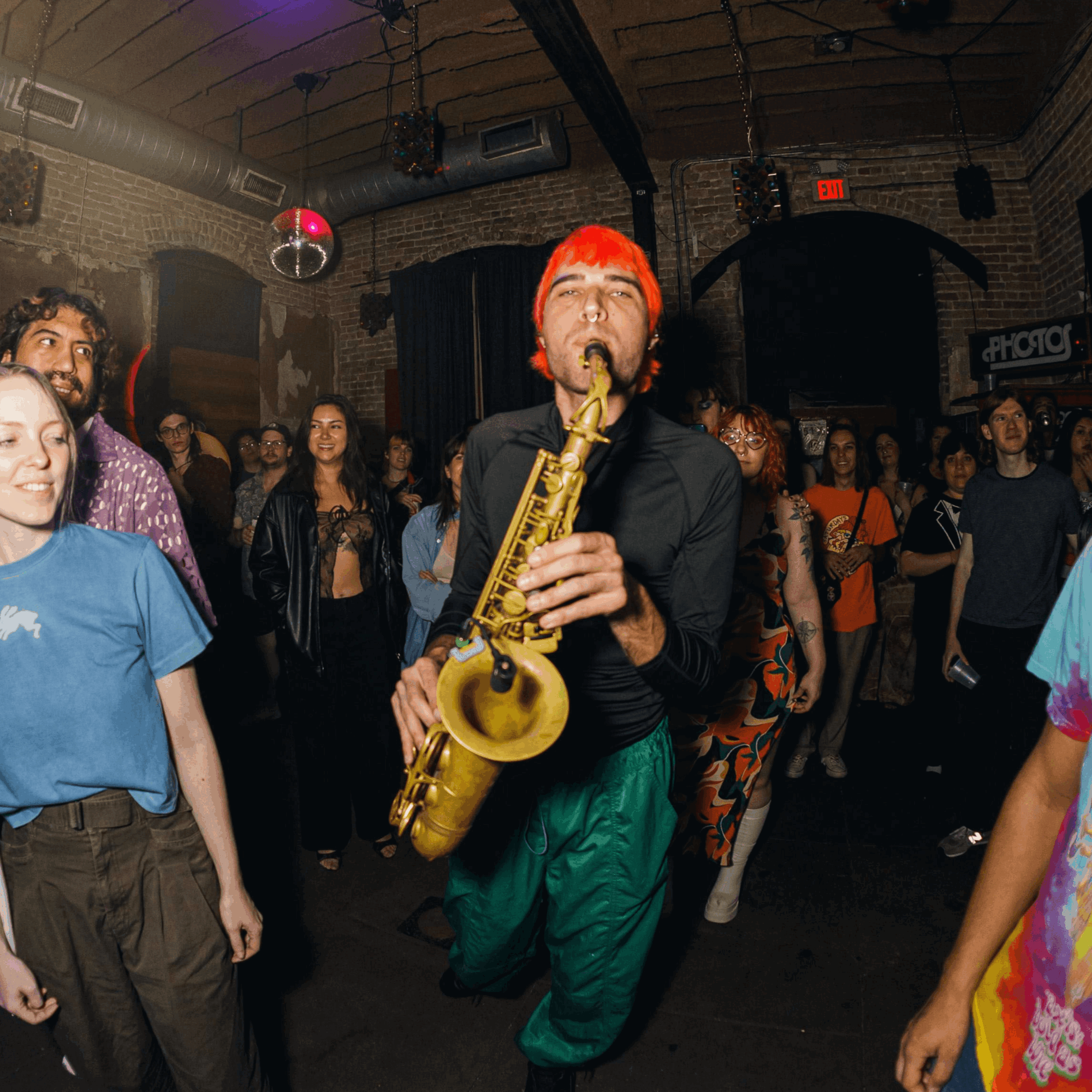 Person with bright orange hair playing saxophone in a lively indoor party with people dancing and socializing.