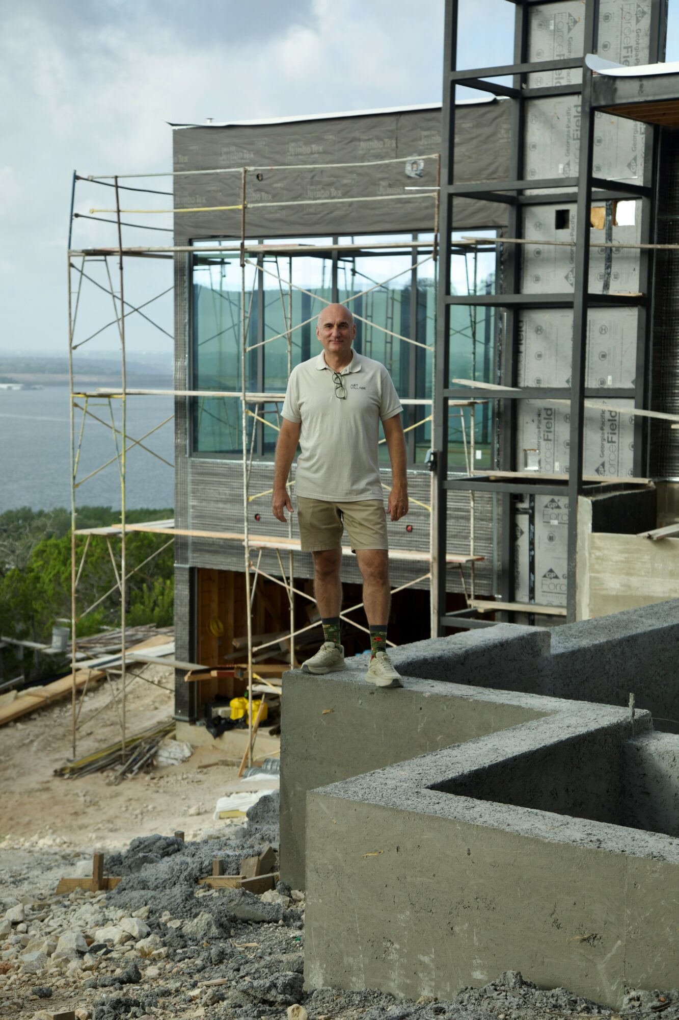 Man standing on a construction site with scaffolding and building materials, overlooking a landscape with water and sky in the background.