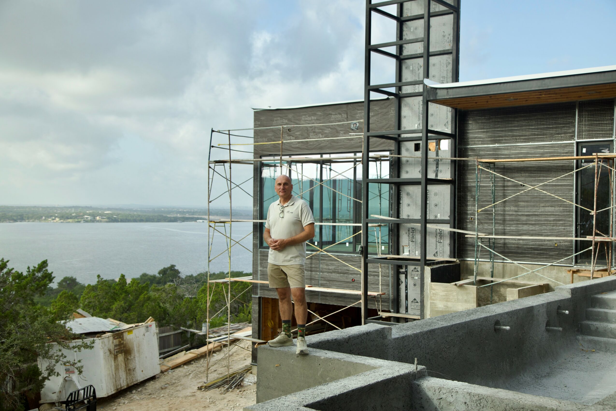 Man standing on a construction site with scaffolding, overlooking a river and landscape in the background.