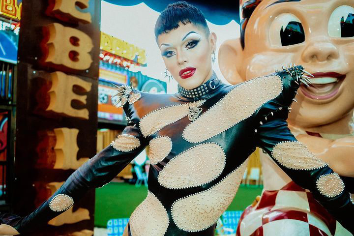 Woman with short dark hair and makeup, wearing a patterned dress, posing with arms outstretched at an amusement park.