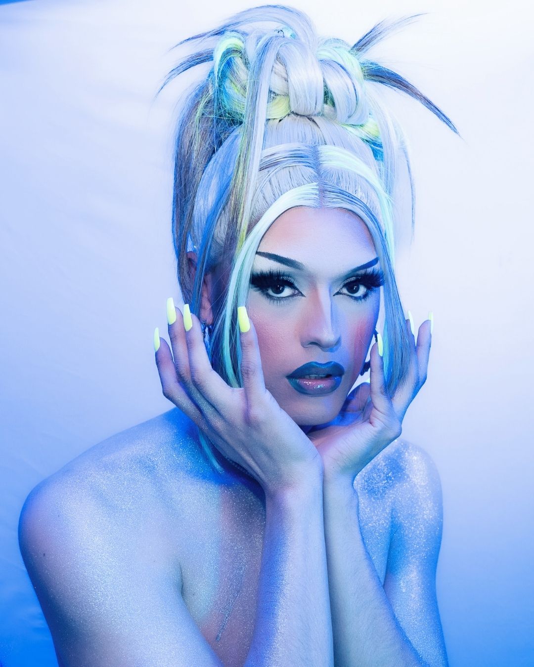 Woman with elaborate pastel-colored hairstyle, bold makeup, and painted blue skin, posing with hands near face against a light background.