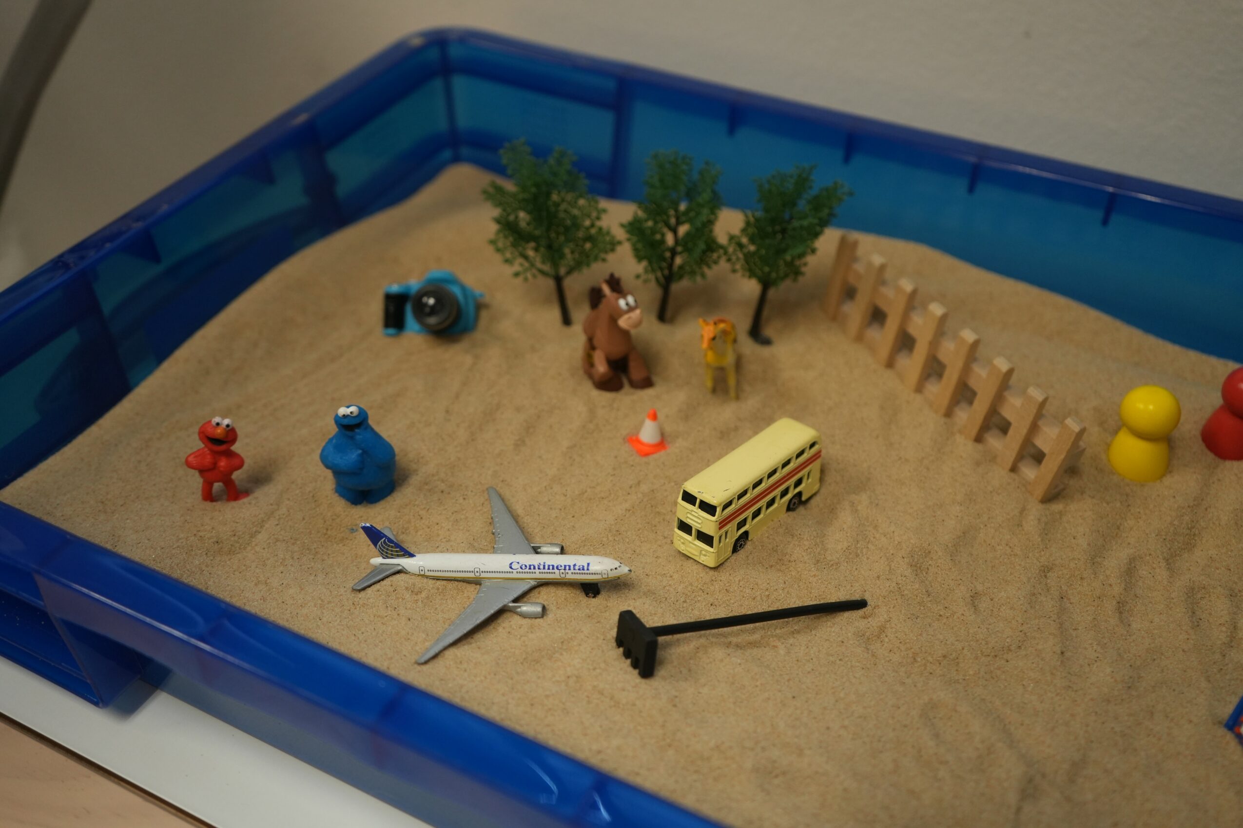 Toy scene with trees, animals, a bus, airplane, and other objects on sand inside a blue container.