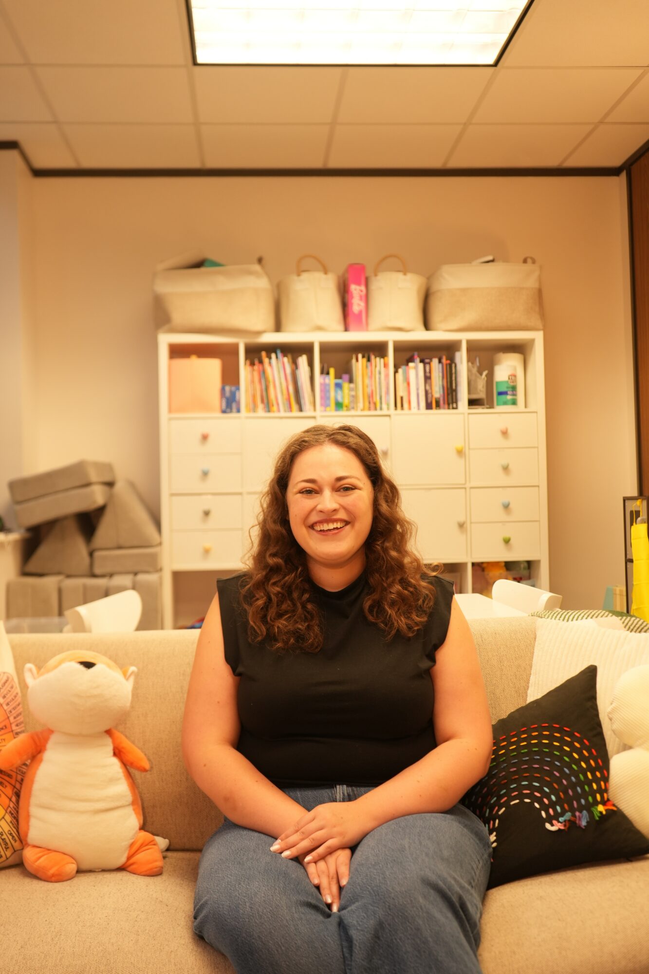 Woman with curly hair smiling, sitting on a beige couch in a room with shelves and stuffed animals.
