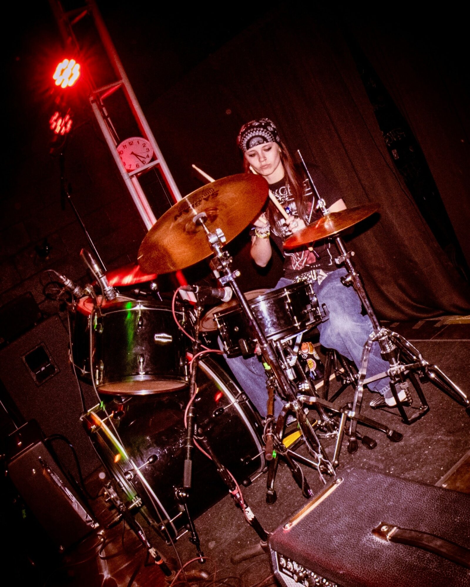 A woman playing drums on stage with red stage lights, wearing a headband and casual clothing.