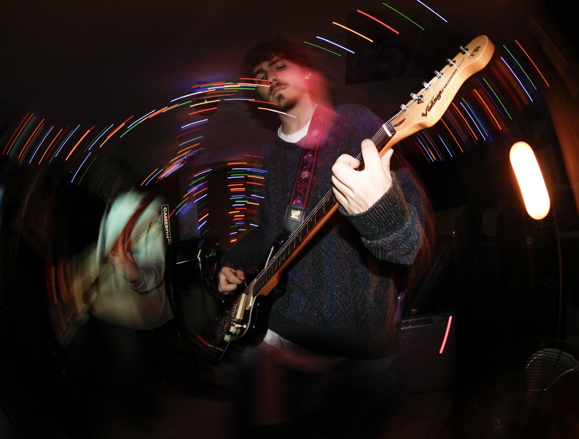 Person playing electric guitar in a dimly lit space with colorful light streaks, motion blur, and a blurred background.