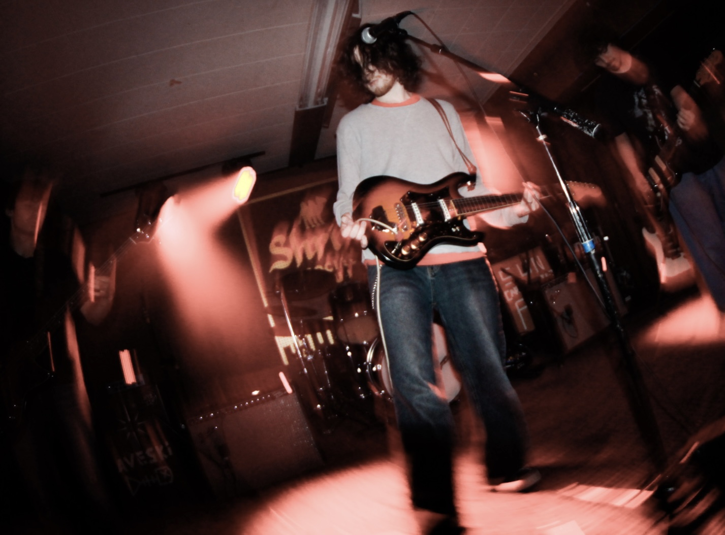 Person playing guitar on stage with pink and red lighting, blurred motion, and a microphone stand nearby.
