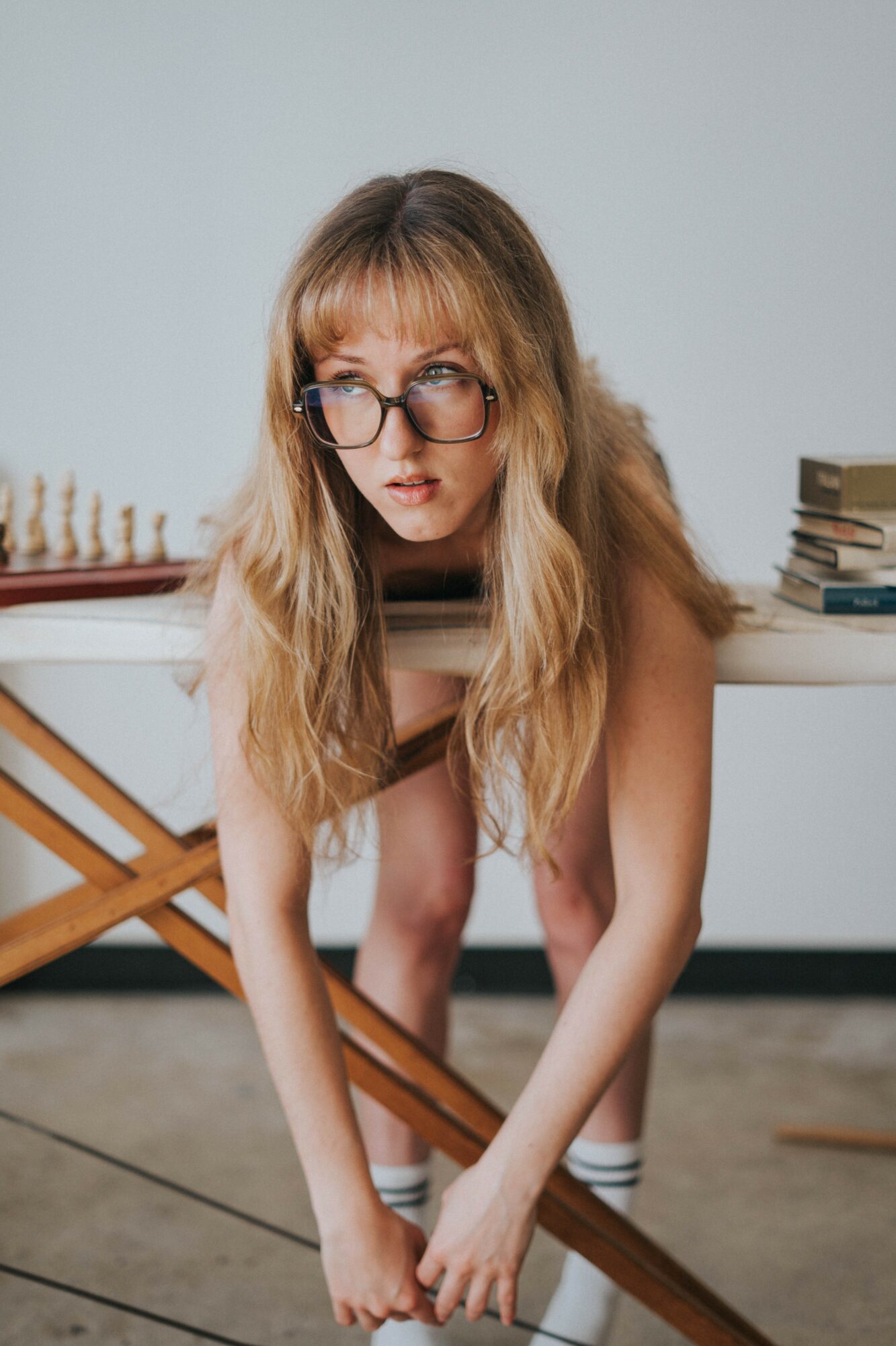 Young woman with glasses and long hair leaning forward, holding a wooden object, with books on a table behind her.