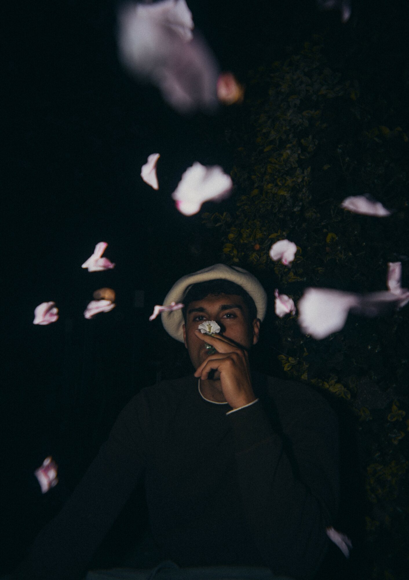 Person wearing a hat and glasses, surrounded by falling pink flower petals at night.