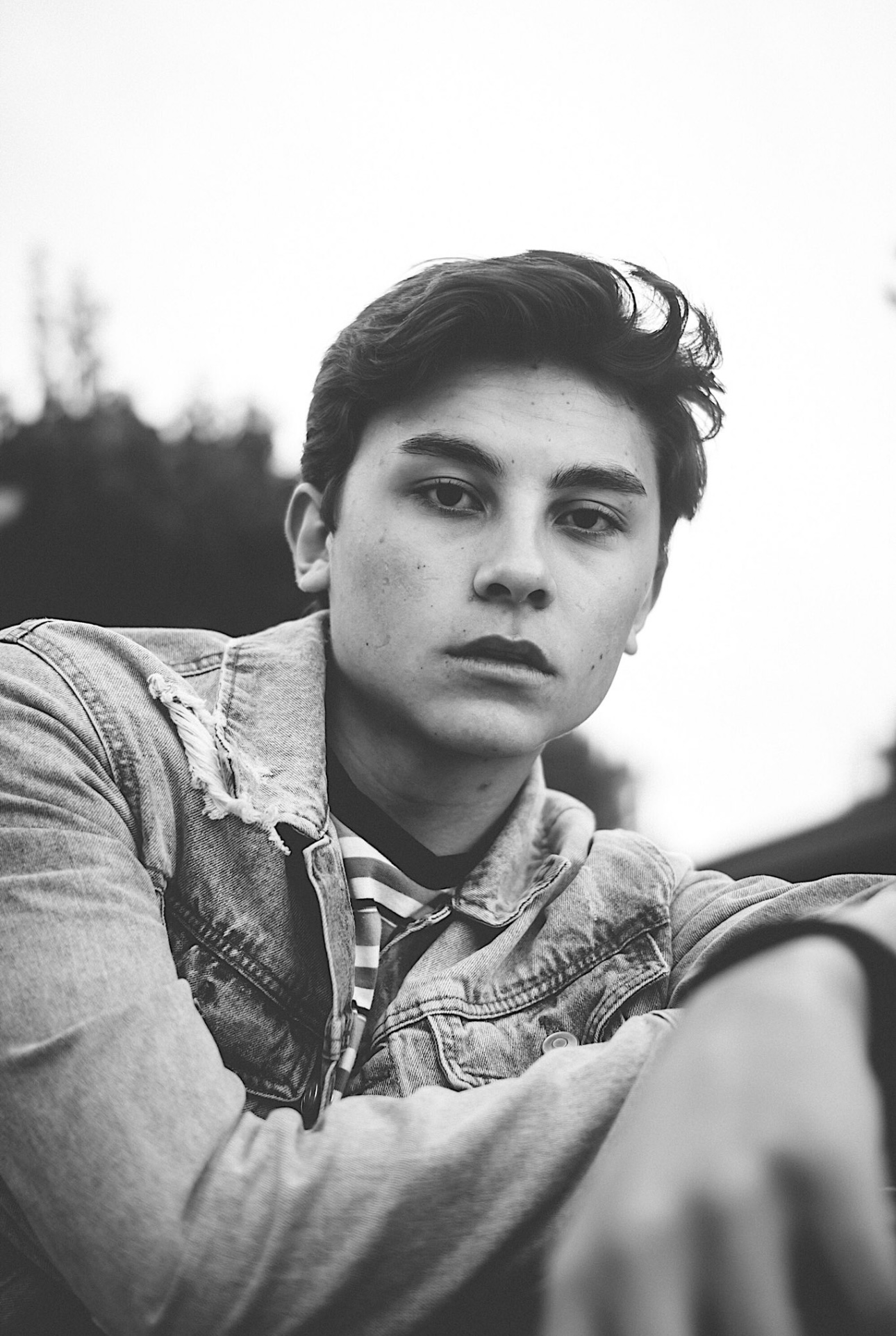 Young person with short dark hair and serious expression, wearing a denim jacket and striped shirt, outdoors.