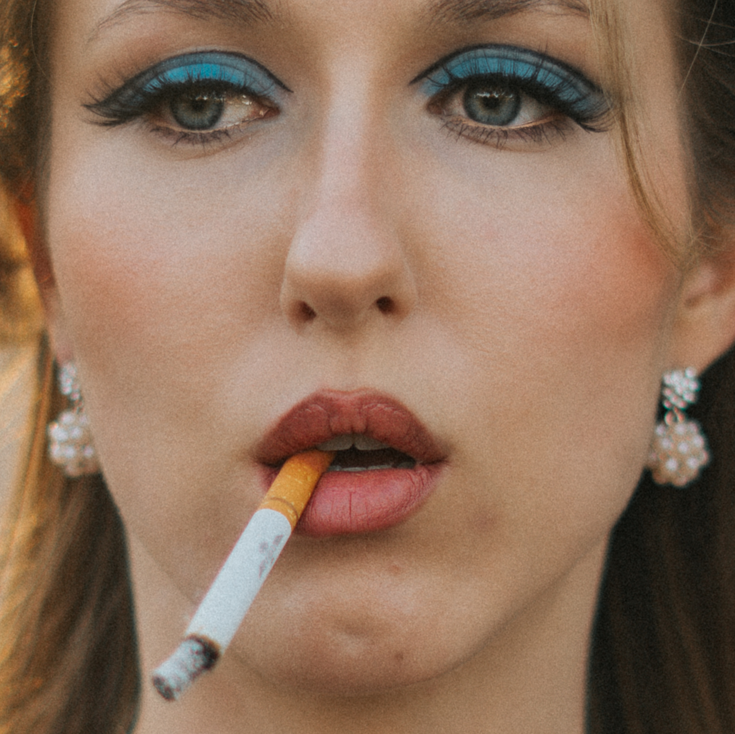 Close-up of a woman with blue eye makeup and earrings, holding a cigarette near her lips.
