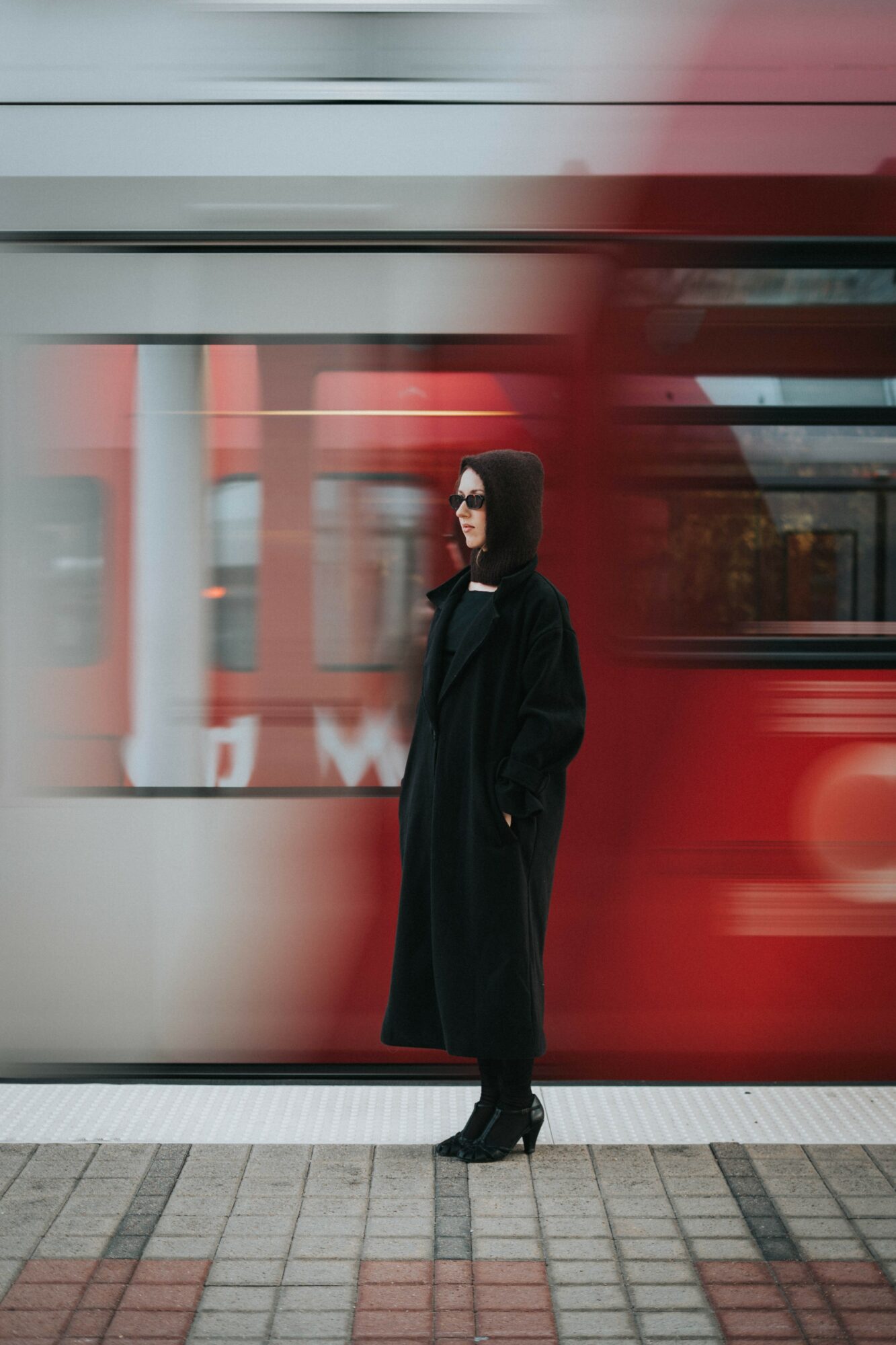 Woman in black coat and sunglasses standing on sidewalk with a moving red bus behind her.