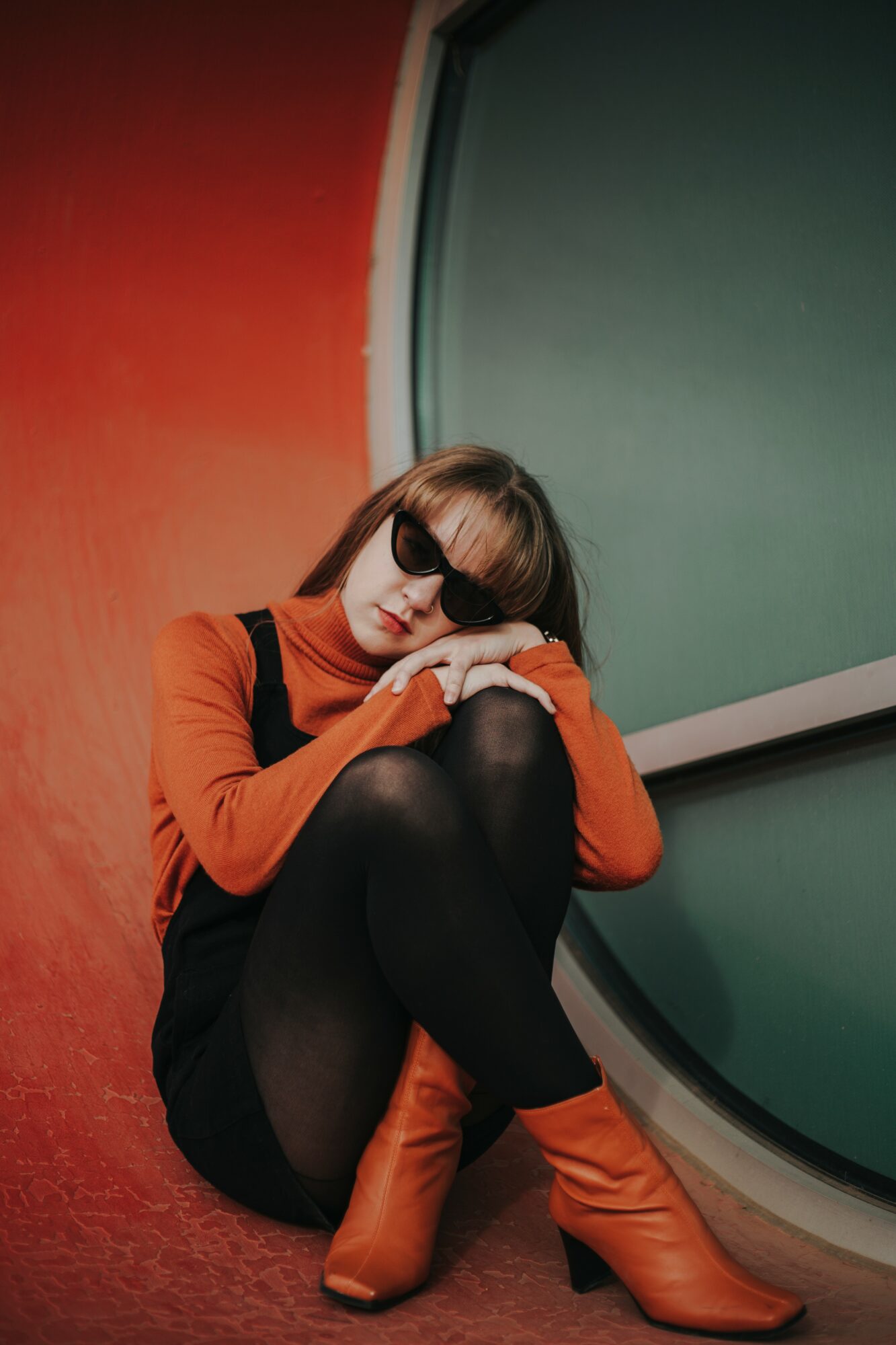 Person sitting on the floor with legs crossed, wearing sunglasses, orange top, black skirt, and orange boots, leaning against a wall.