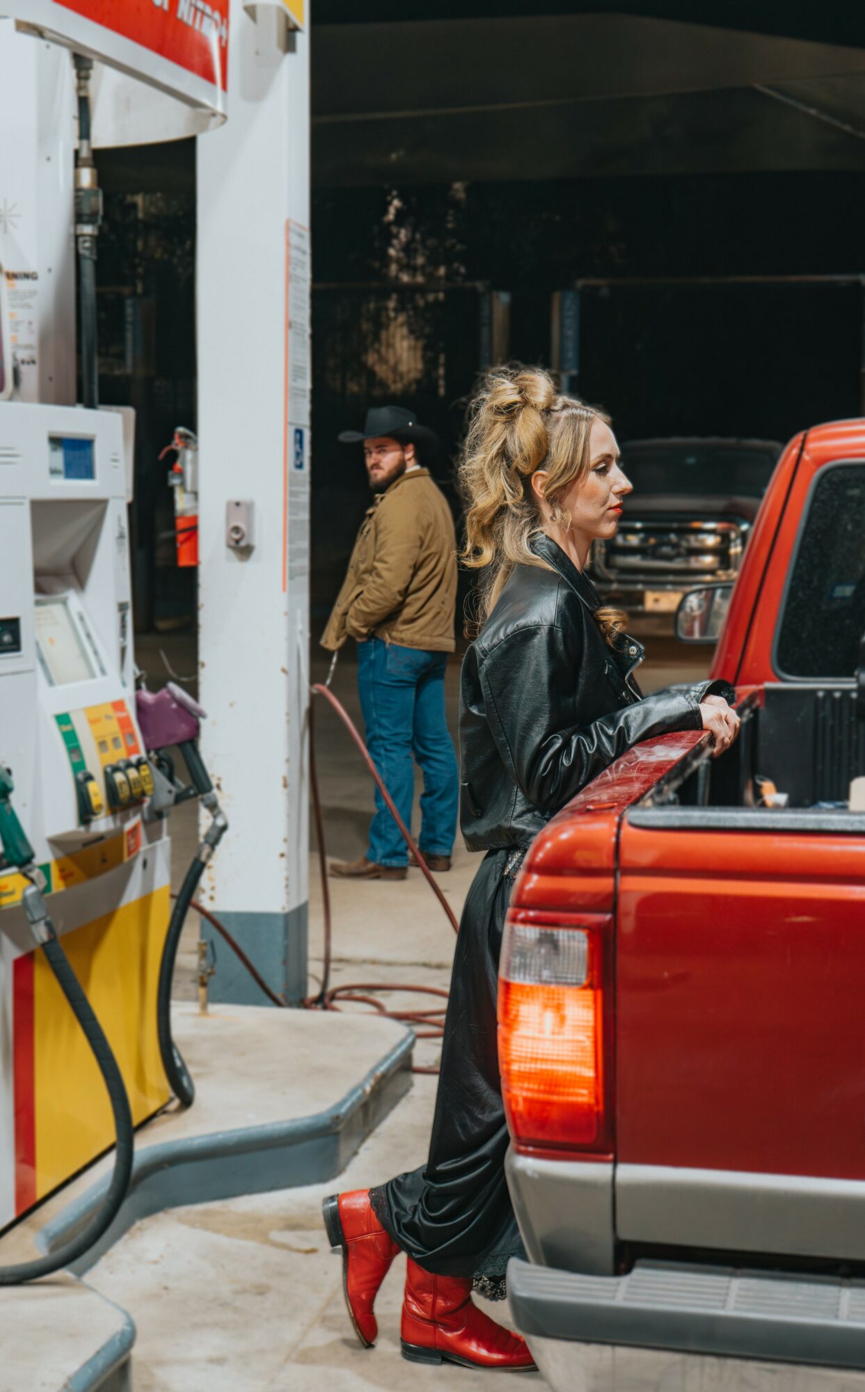 Woman with blonde hair in a ponytail, wearing a black leather jacket and red boots, filling gas at a station.