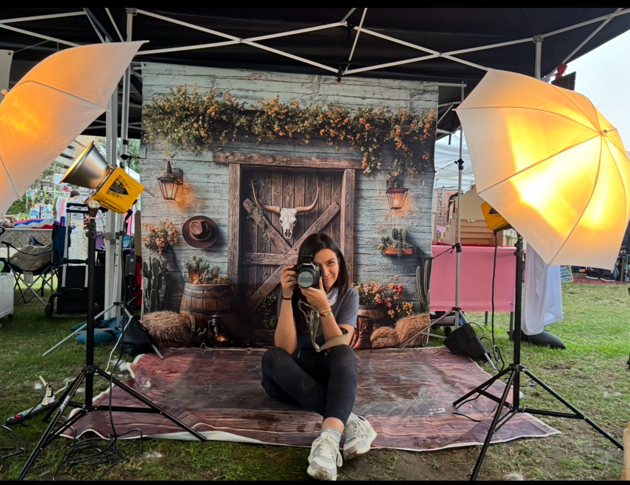 Person sitting on a rug, taking a photo in front of a rustic backdrop with umbrellas and lighting equipment.